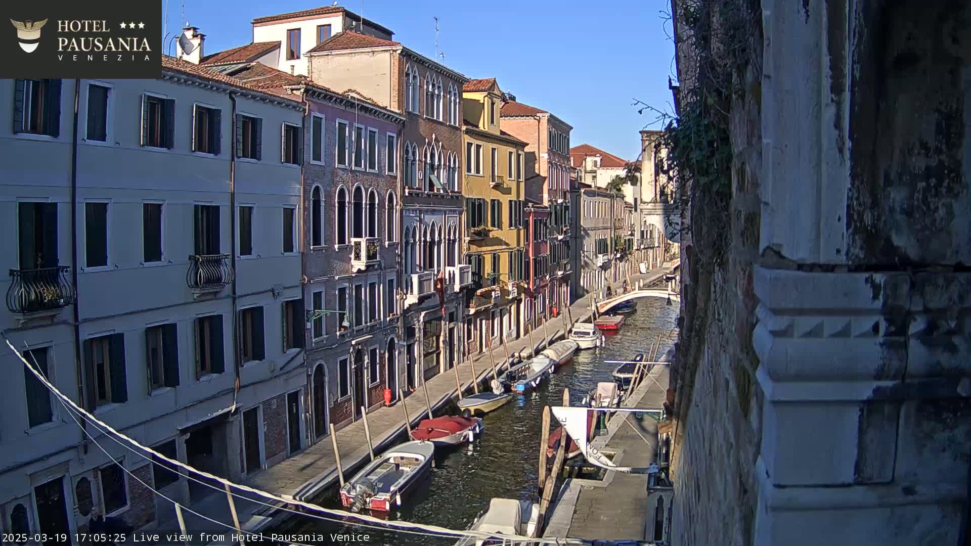 A sunny day in Venice shows several boats moored along a canal, with colorful buildings lining the waterway.