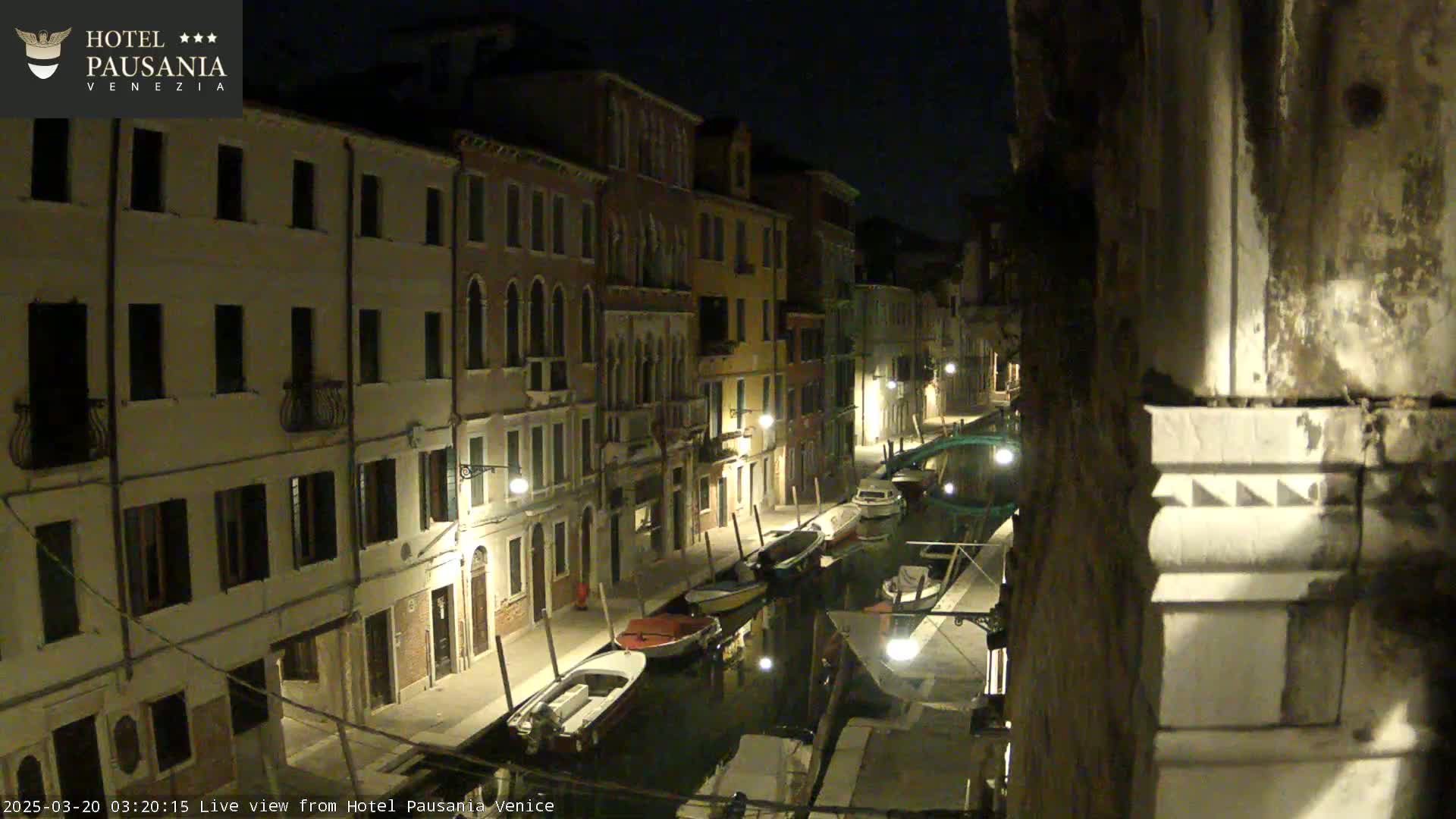 A nighttime view of a narrow canal in Venice, Italy, lined with buildings and several moored boats.