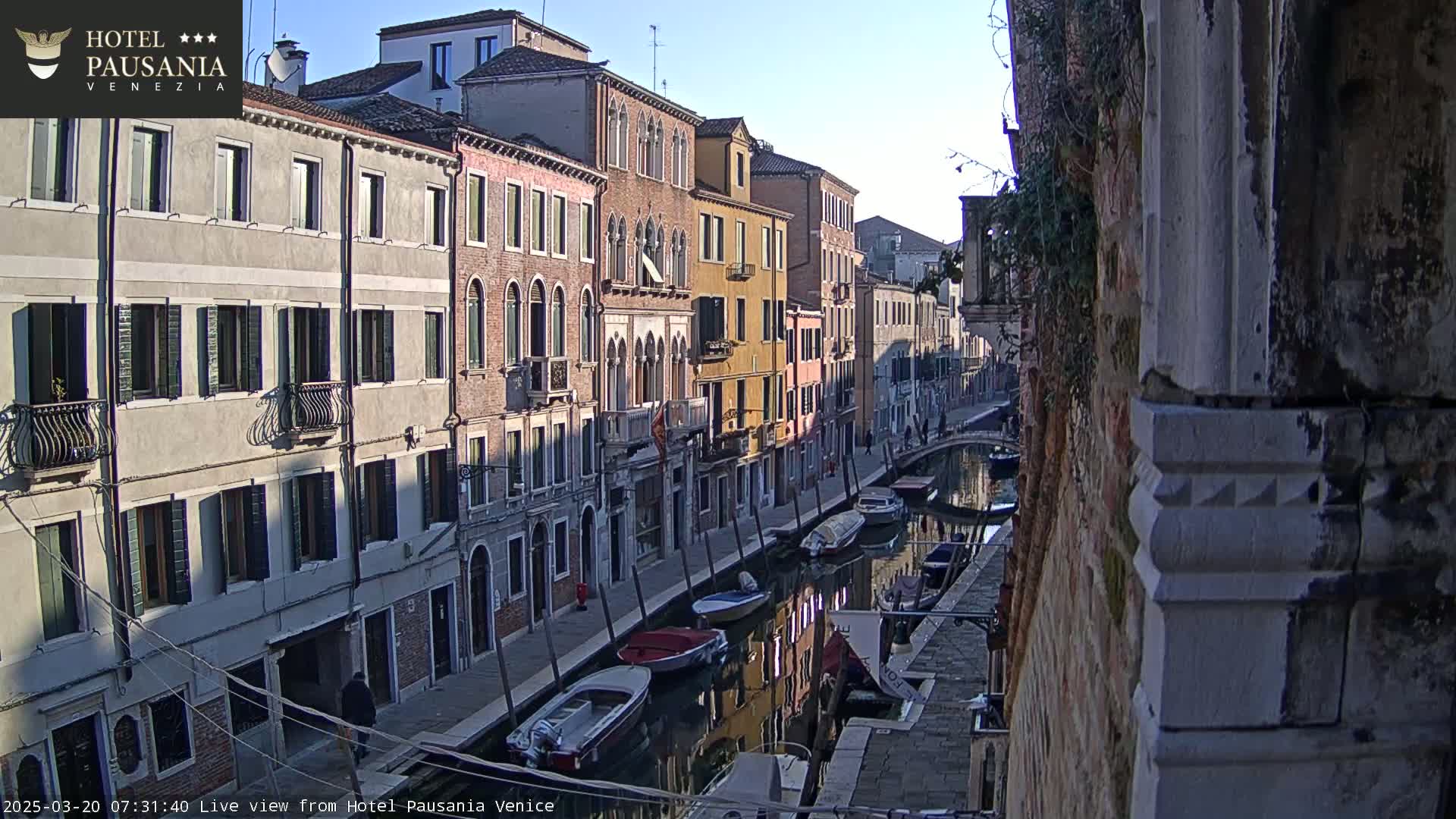 A sunny morning view of a narrow canal in Venice, lined with tall, closely-packed buildings and several small boats moored along its banks.