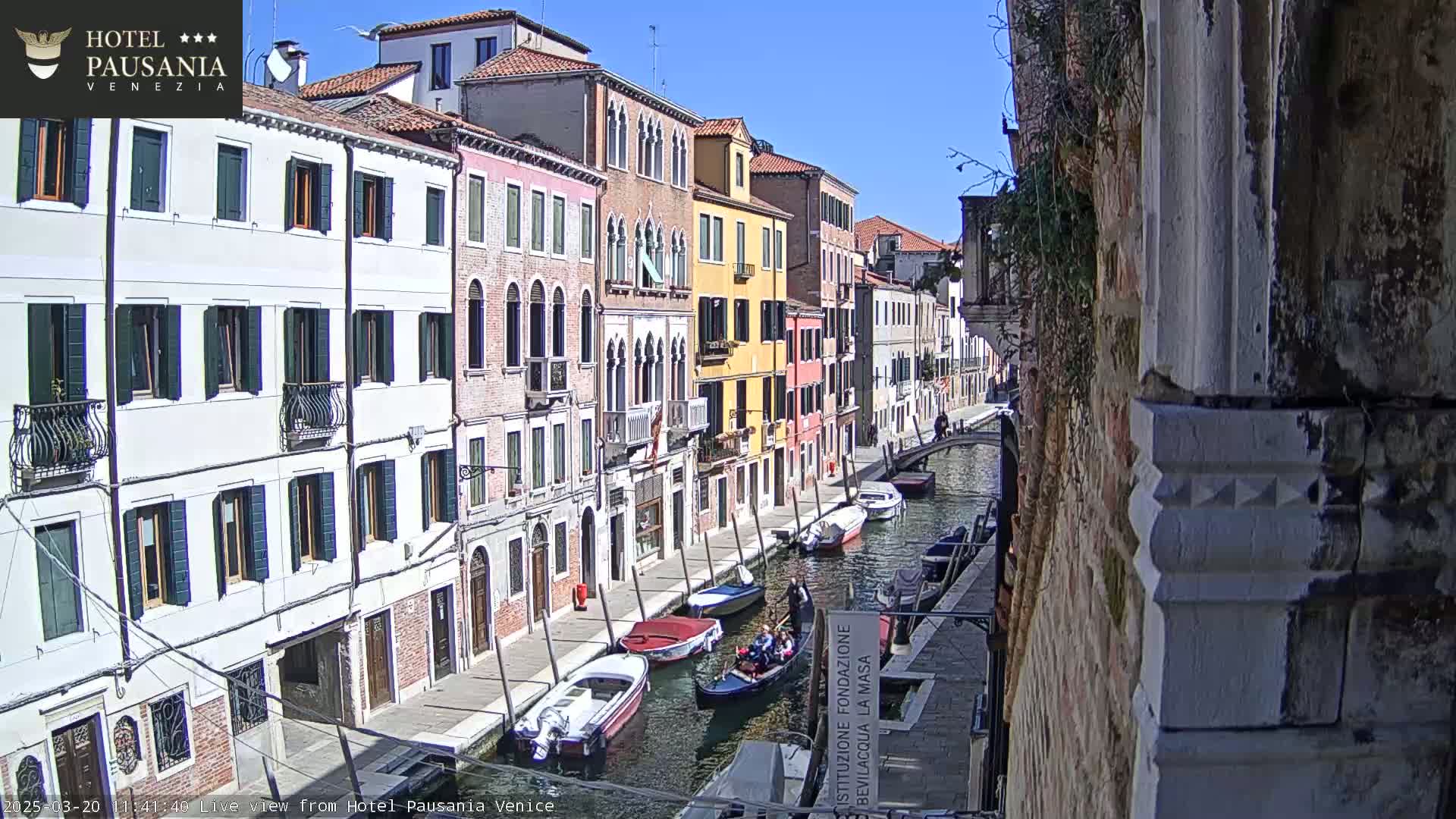 Under a clear blue sky, several boats are moored along a narrow canal lined with colorful, historic buildings.