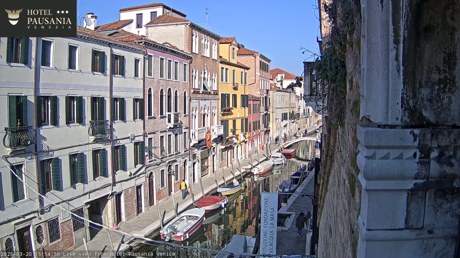 A sunny day in Venice shows a narrow canal lined with colorful buildings, several small boats moored along the waterway, and a few pedestrians walking on the adjacent sidewalk.