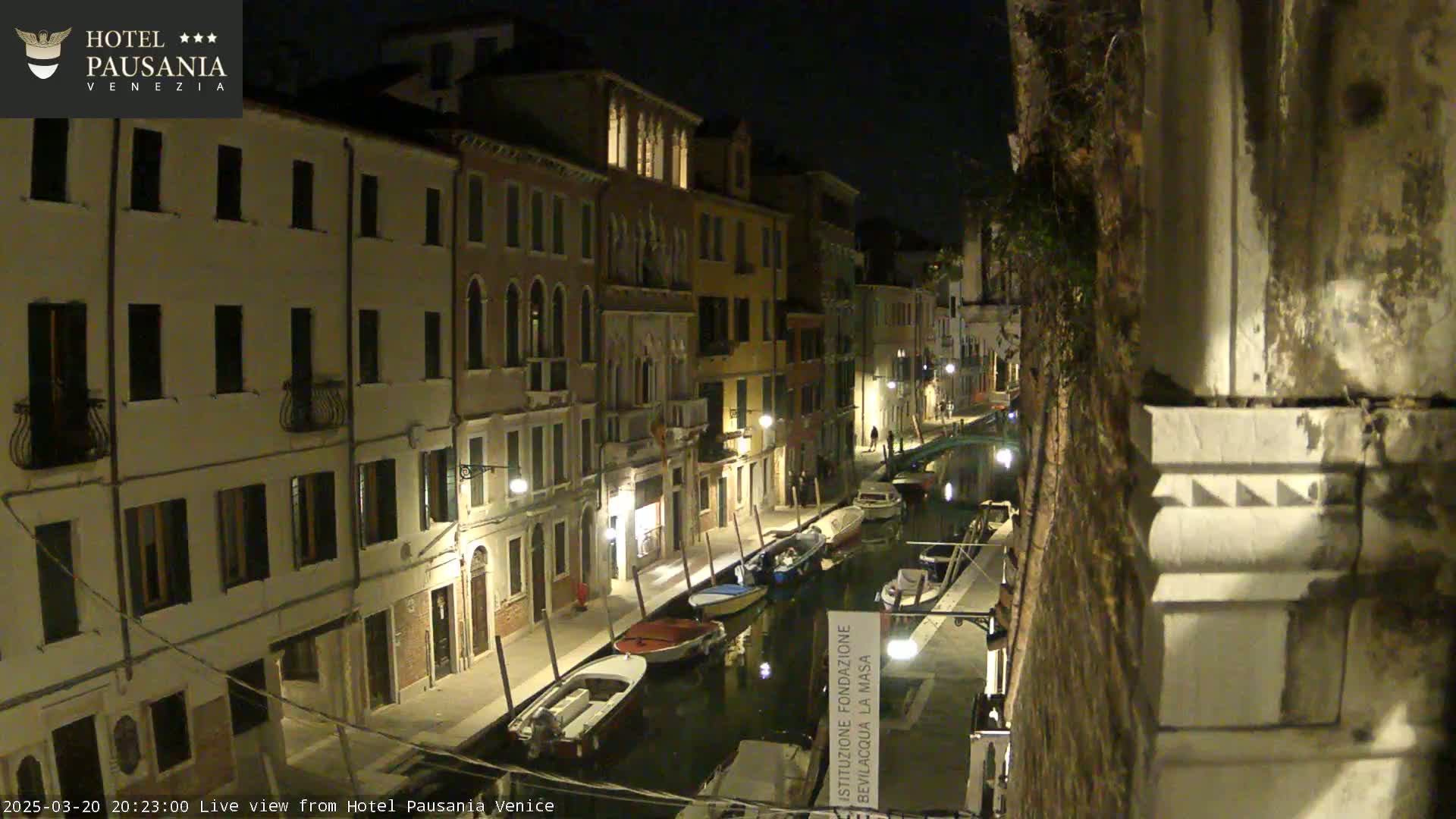 A nighttime view of a narrow canal in Venice, Italy, lined with illuminated buildings and several moored boats.