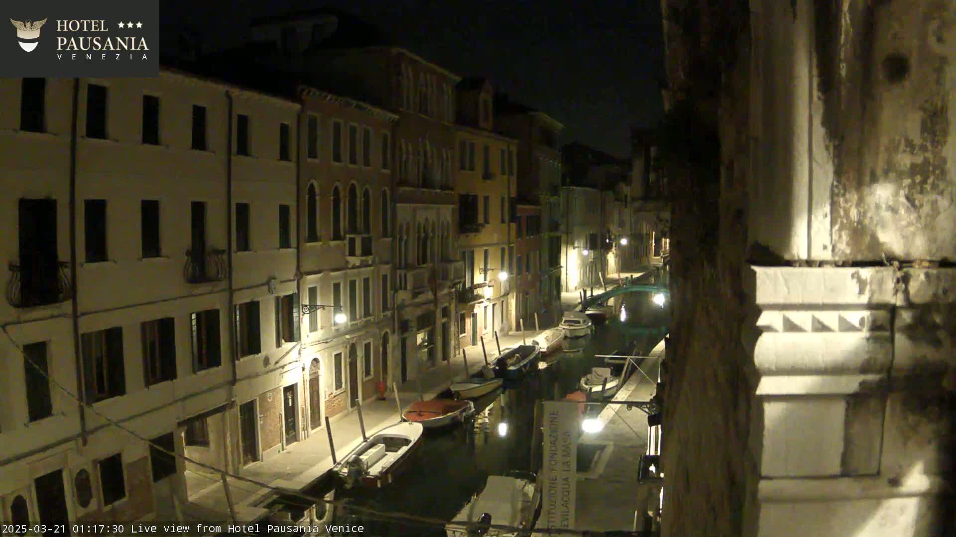 A nighttime view of a canal in Venice, showing several boats moored alongside buildings lit by streetlights.
