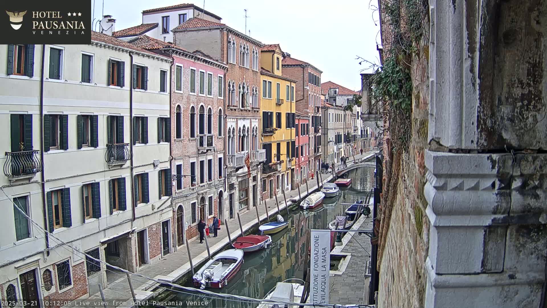 A canal in Venice, with colorful buildings lining the waterway, boats moored along the sides, and an overcast sky.