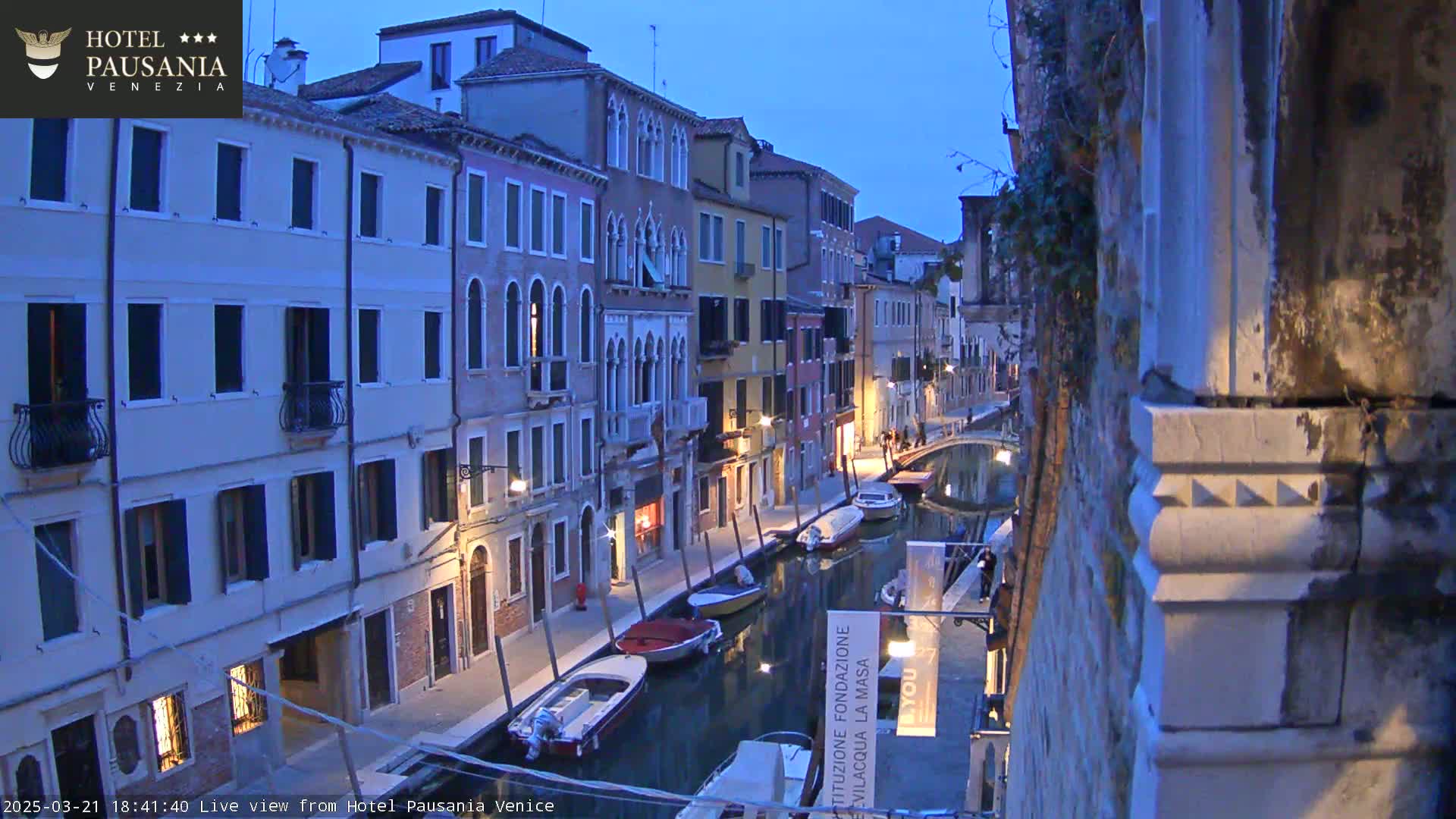 A twilight view of a narrow canal in Venice, lined with closely spaced buildings, with several small boats moored along its sides.