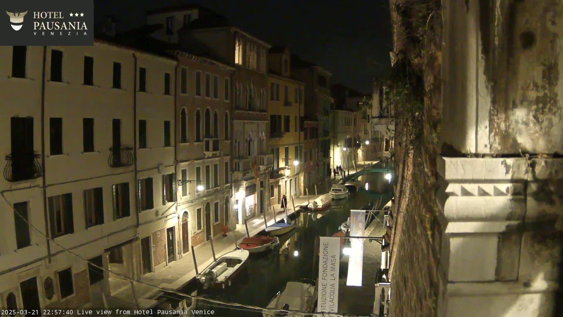 A nighttime view of a canal in Venice, Italy, with boats moored along the sides of buildings illuminated by streetlights.
