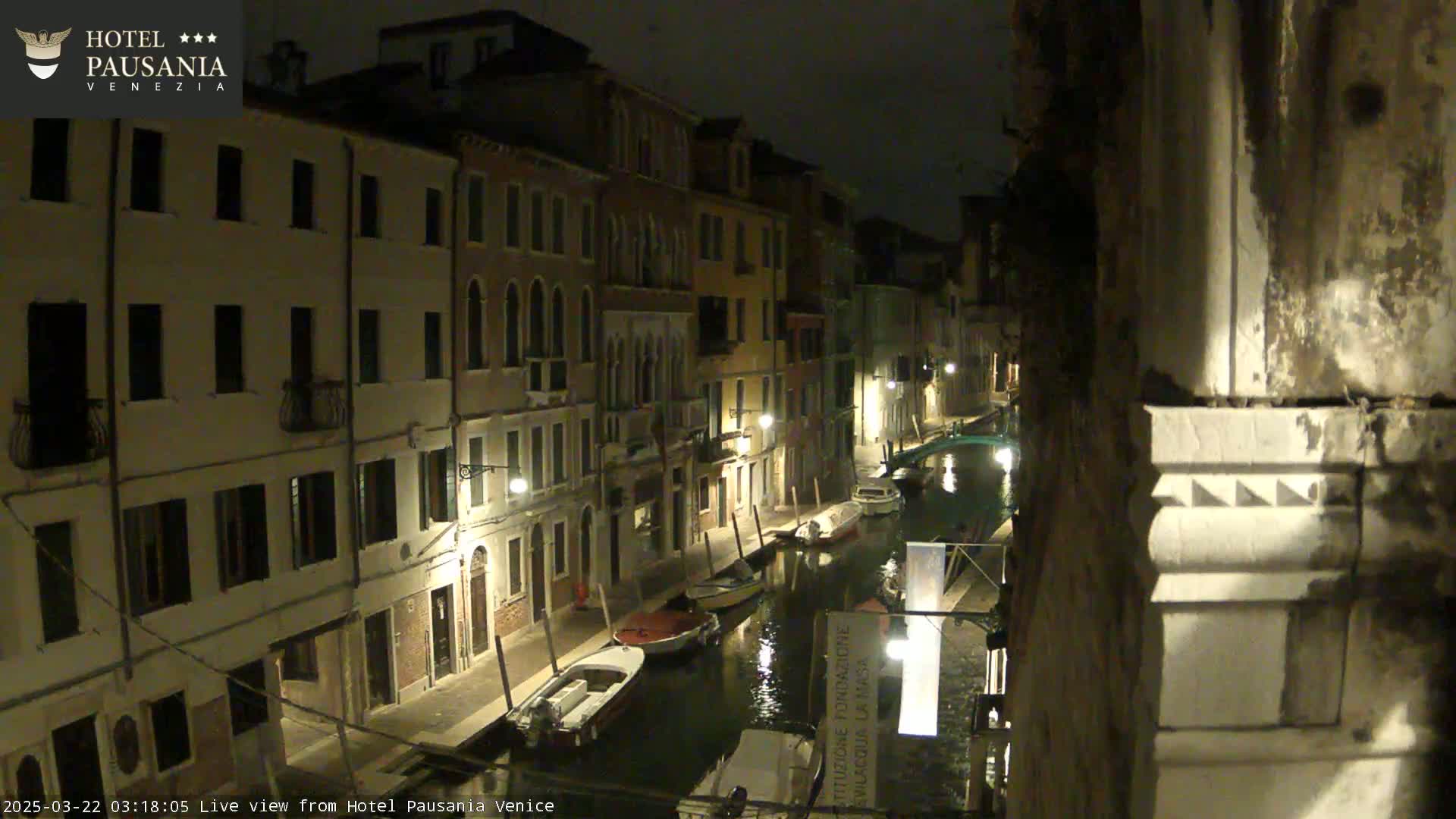 A nighttime view of a canal in Venice, showing several boats moored along the waterway and buildings lining the sides, illuminated by streetlights.