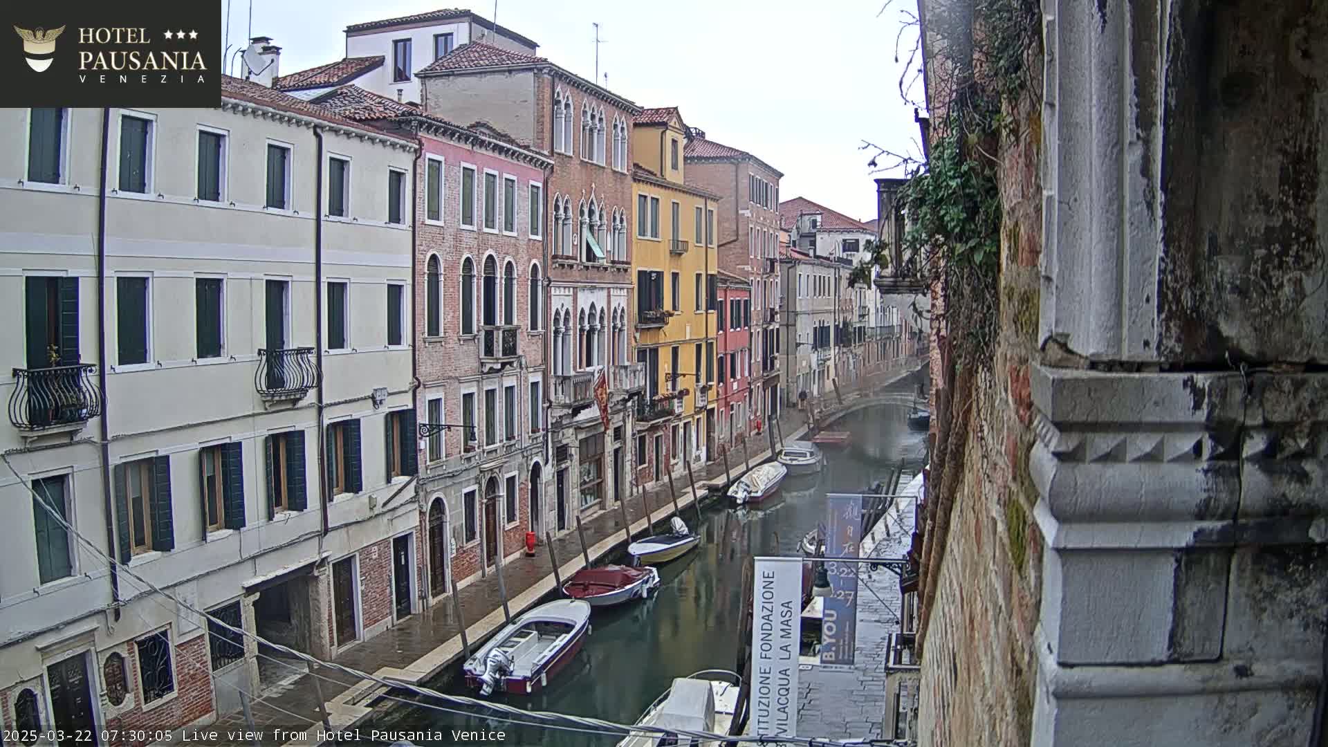 A narrow canal in Venice, under an overcast sky, is lined with closely-spaced buildings and several small boats moored along its edges.