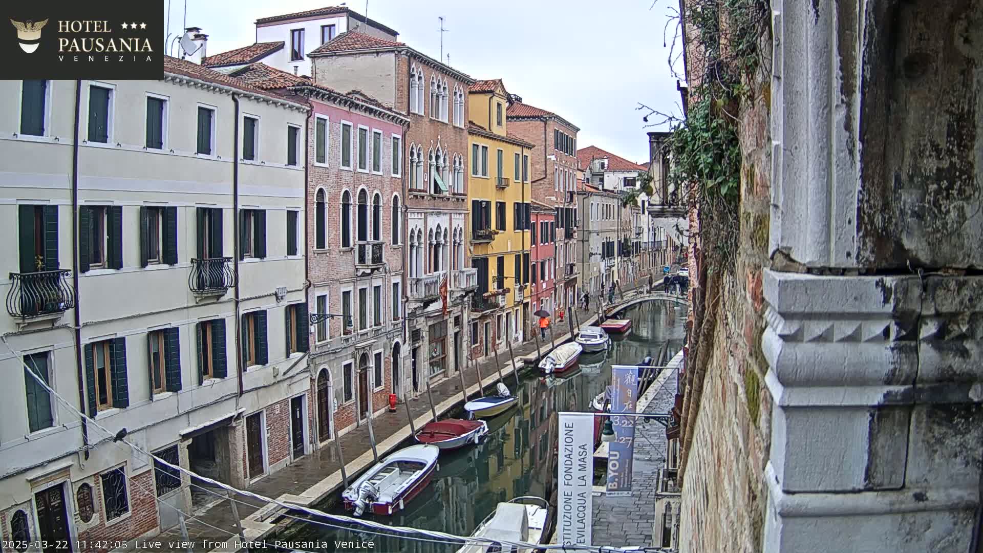 A view down a narrow Venetian canal on an overcast day shows several moored boats and colorful buildings lining the waterway.