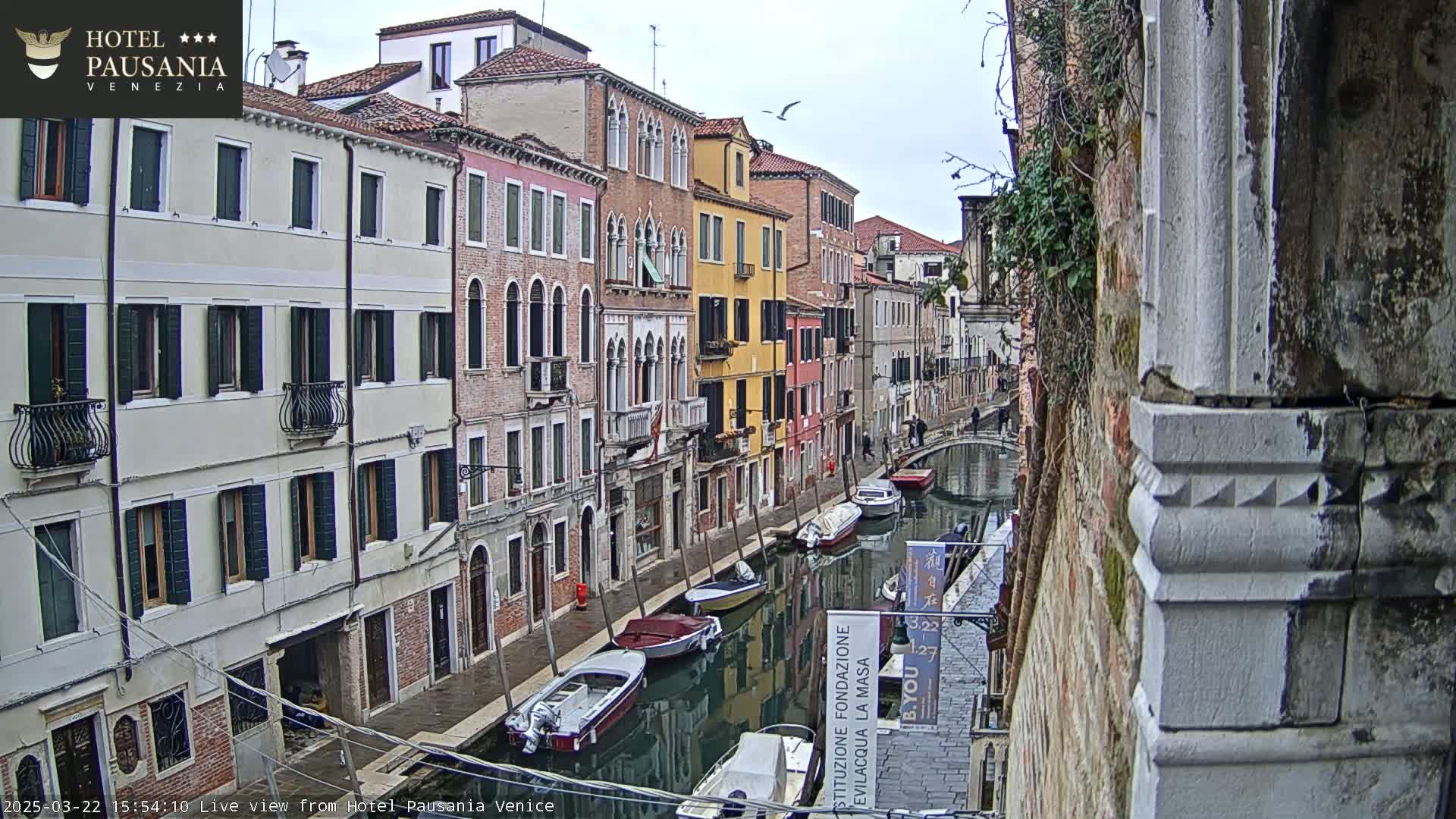 A narrow canal in Venice, Italy, is lined with colorful buildings on a cloudy day, and several boats are moored along its banks.