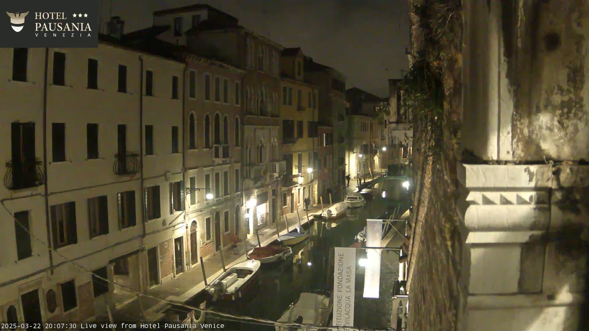 A nighttime view of a narrow canal in Venice, Italy, lined with illuminated buildings and several moored boats.