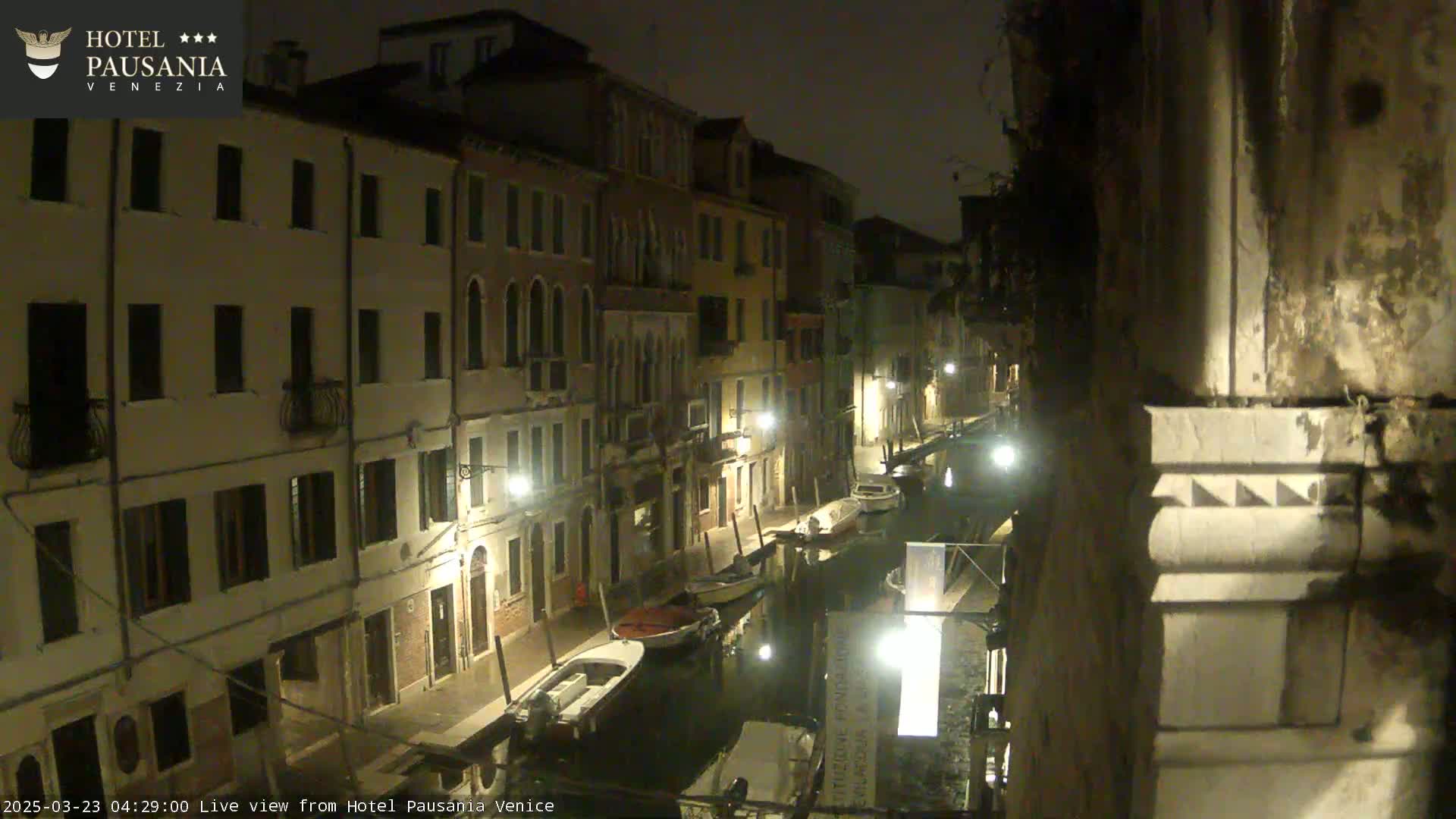 A nighttime view of a narrow canal in Venice, Italy, lined with buildings and a few moored boats, under overcast skies.