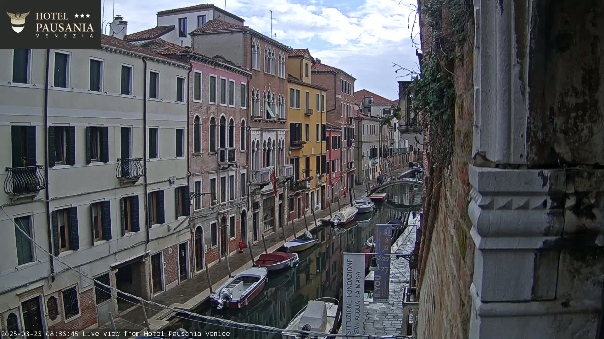 A narrow canal in Venice, under partly cloudy skies, is lined with colorful buildings and several moored boats.