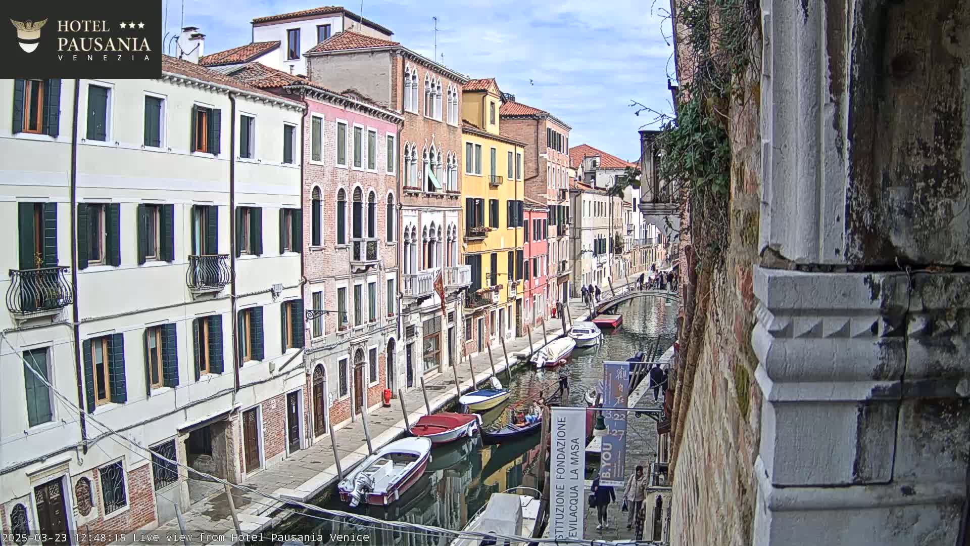 A narrow canal in Venice, Italy, lined with colorful buildings, is filled with several boats on a partly sunny day.