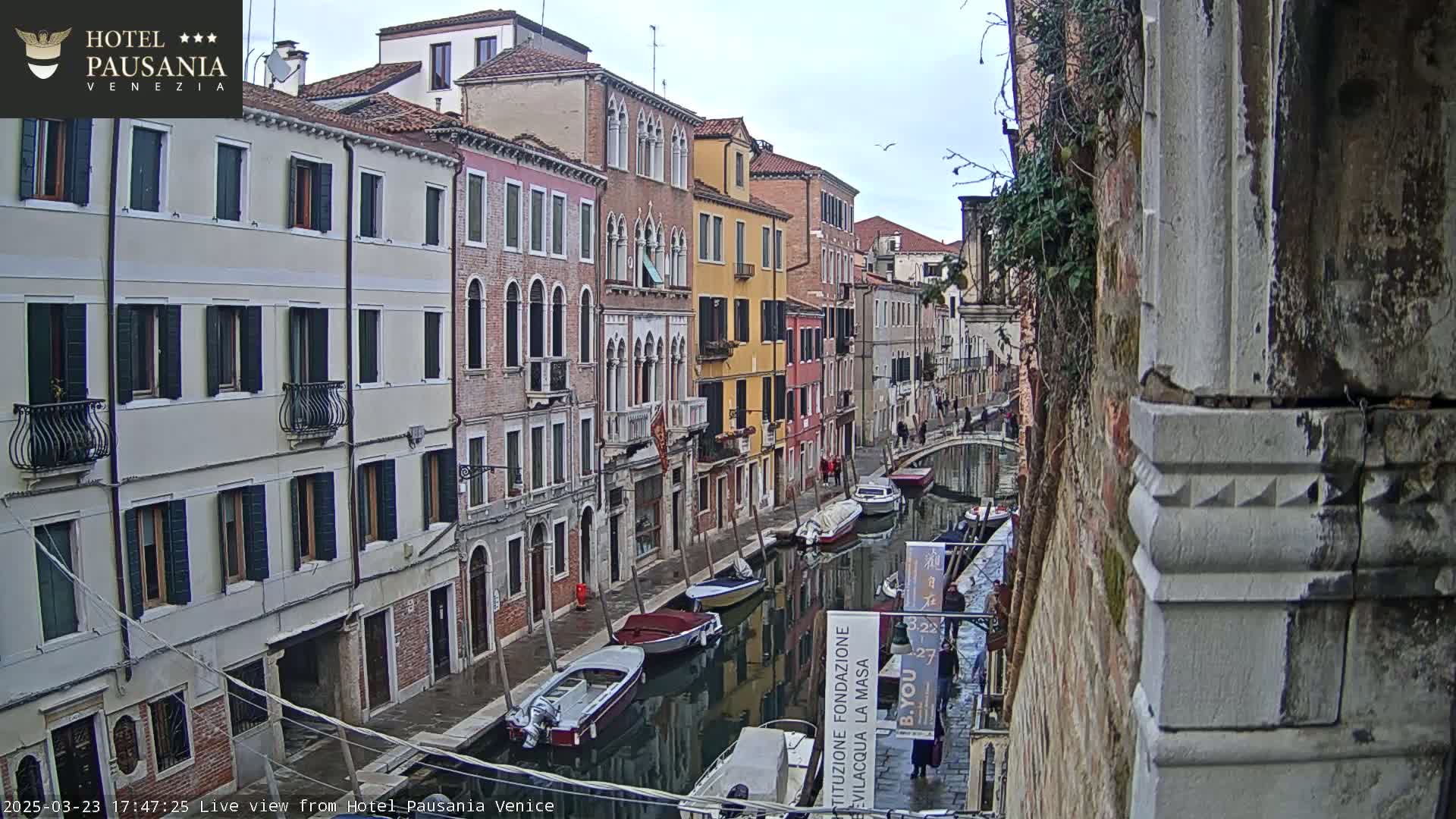 A narrow canal in Venice, Italy, is lined with colorful buildings under an overcast sky; several boats are moored along the canal's edge.