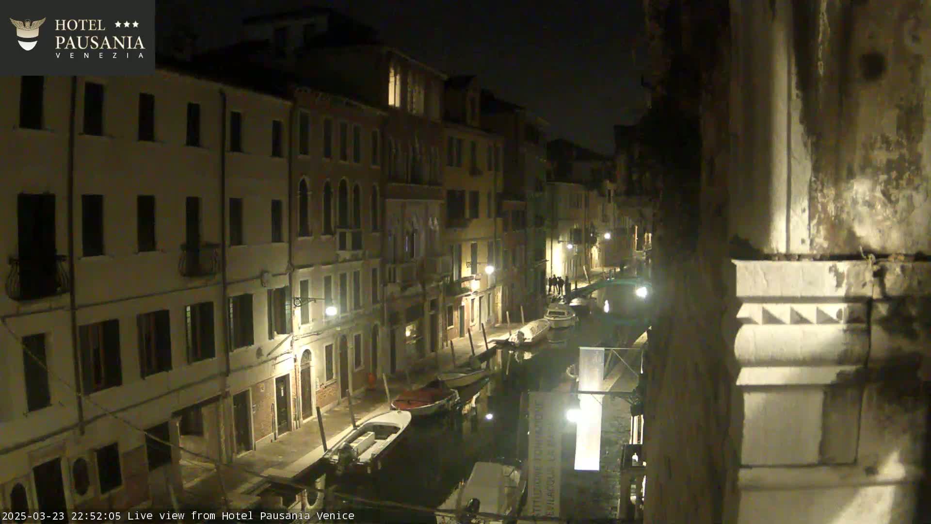 A nighttime view of a narrow canal in Venice, lined with illuminated buildings, with several boats moored along its banks.