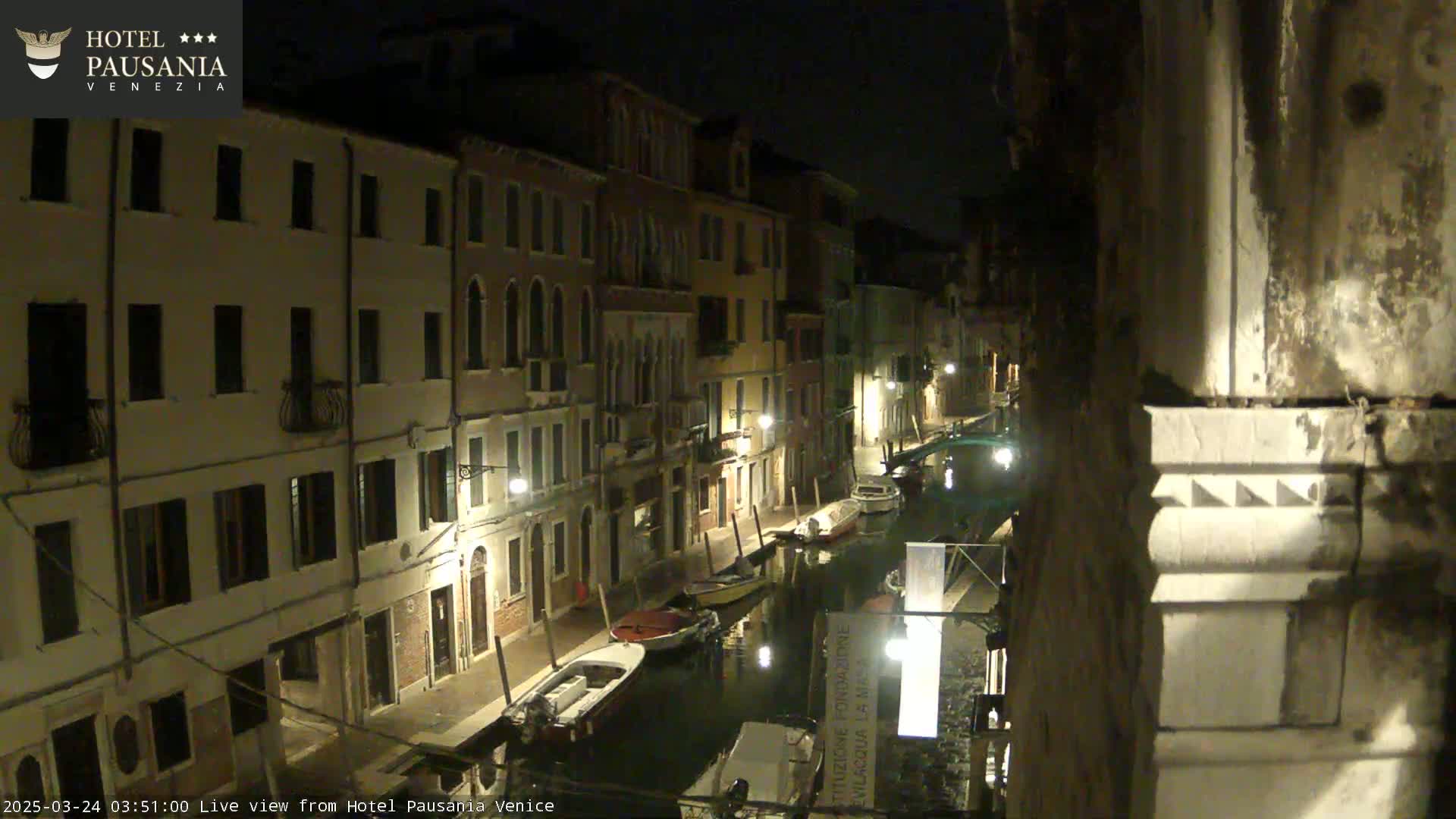 A nighttime view of a narrow canal in Venice, lined with illuminated buildings and several moored boats.