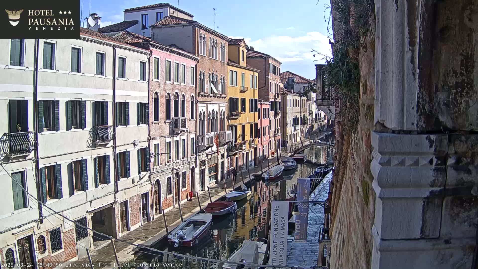 A sunny day in Venice shows a canal lined with colorful buildings and several boats moored along its banks.