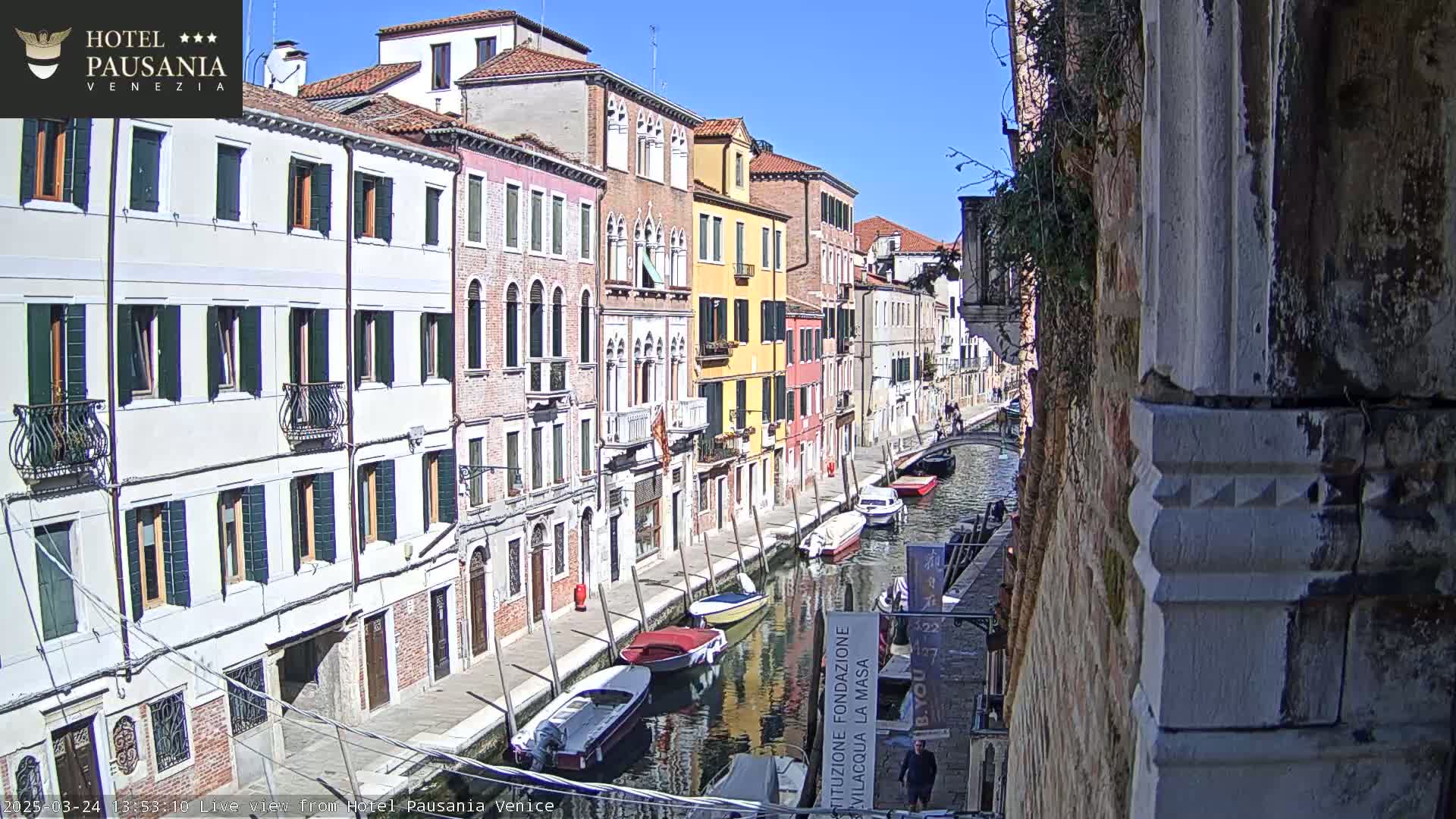 A sunny day in Venice shows a narrow canal lined with colorful buildings, and several boats are moored along its sides.