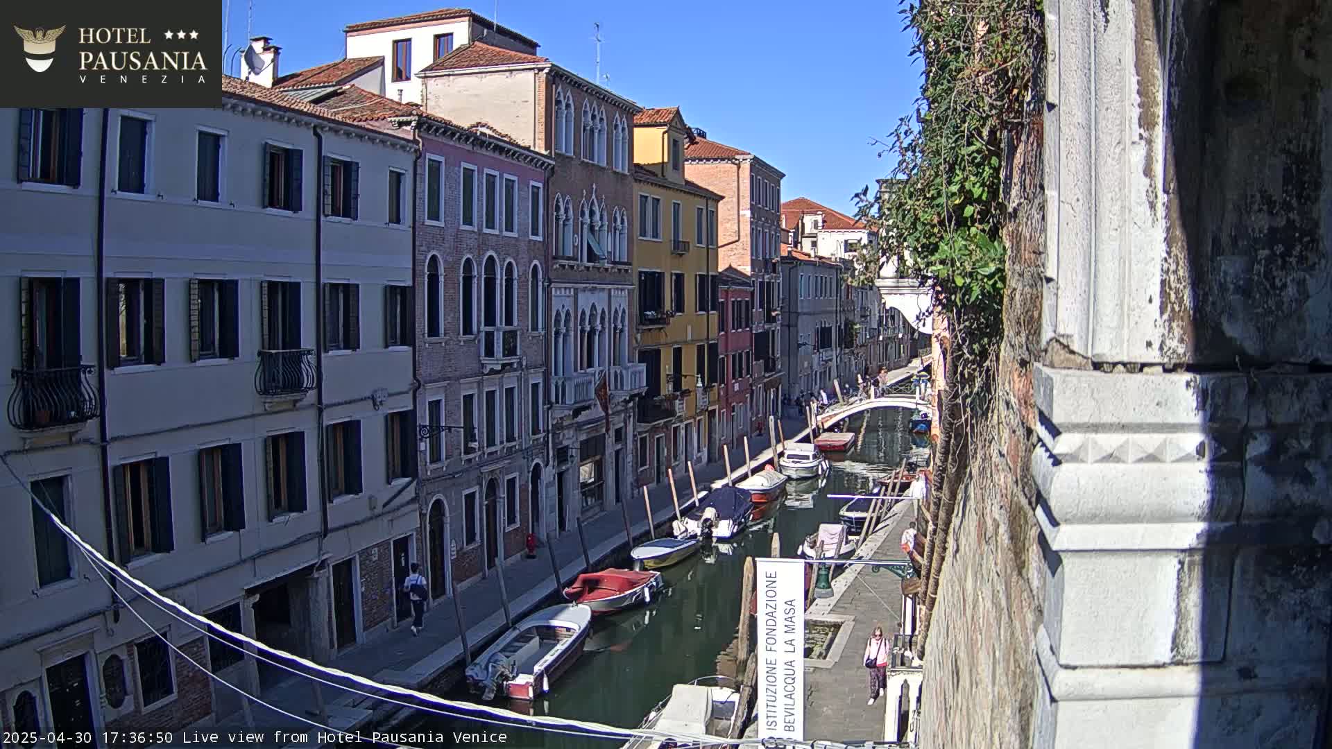 A sunny day in Venice shows a narrow canal lined with colorful buildings and several moored boats.