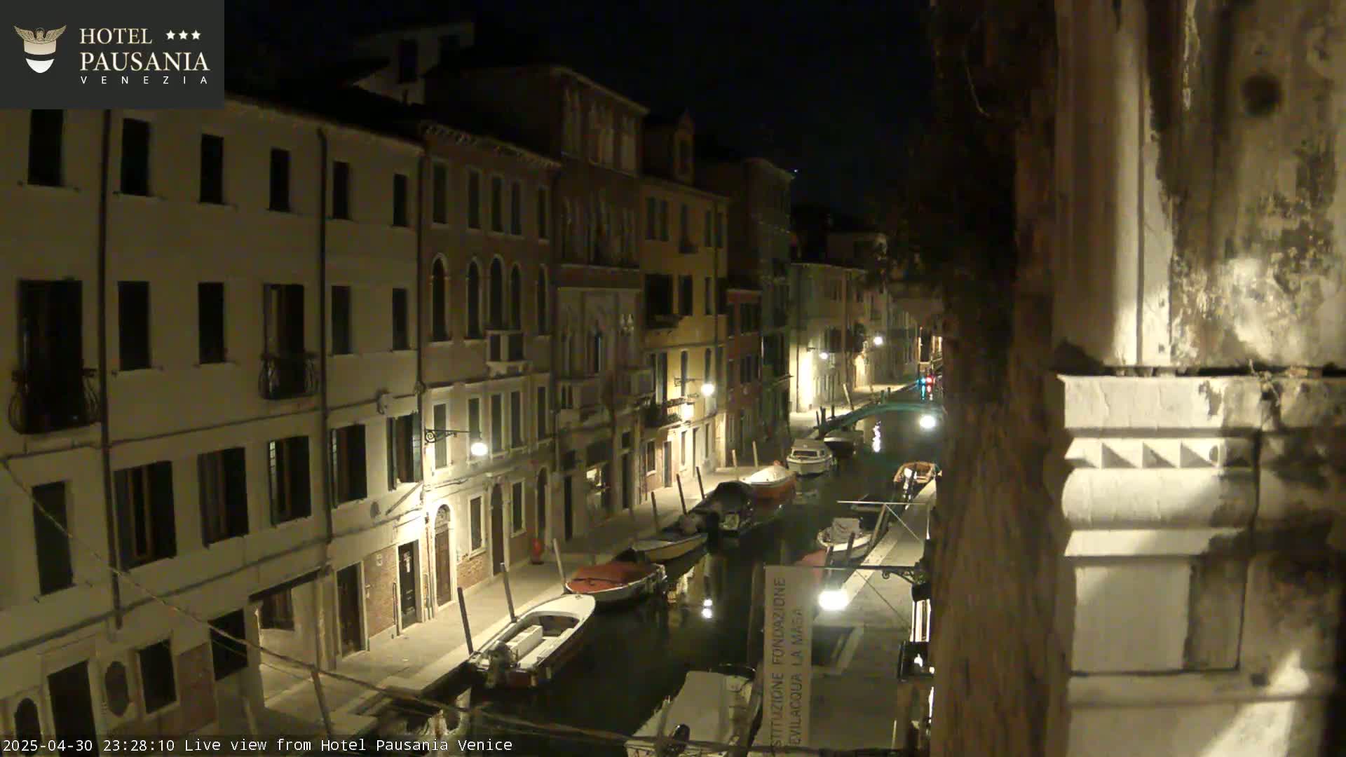 A nighttime view of a narrow canal in Venice, lined with illuminated buildings and several moored boats.