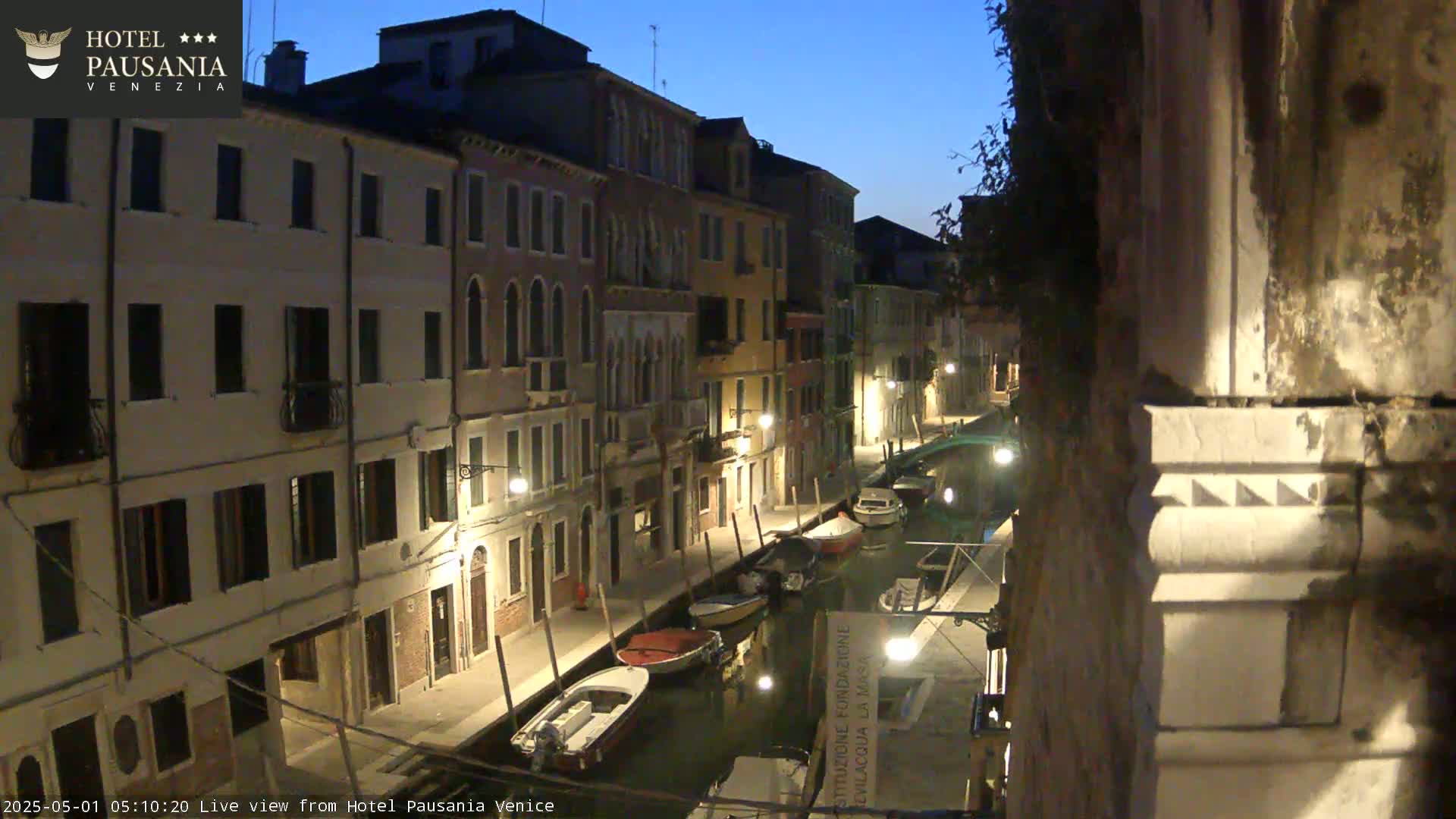 A nighttime view of a narrow canal in Venice, lined with illuminated buildings and several moored boats, under a twilight sky.