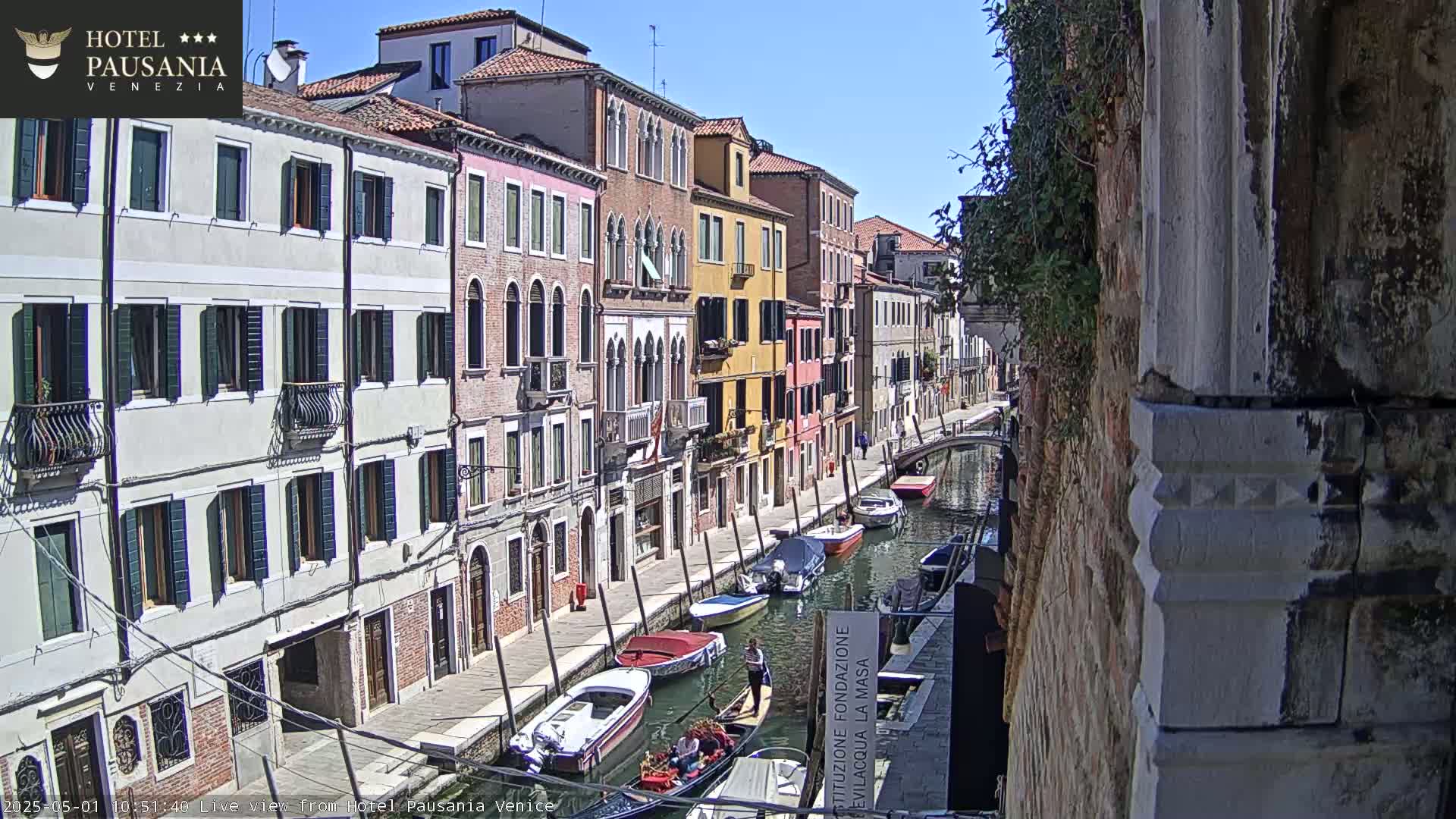 A sunny day in Venice shows a canal lined with colorful buildings and several boats, including gondolas.