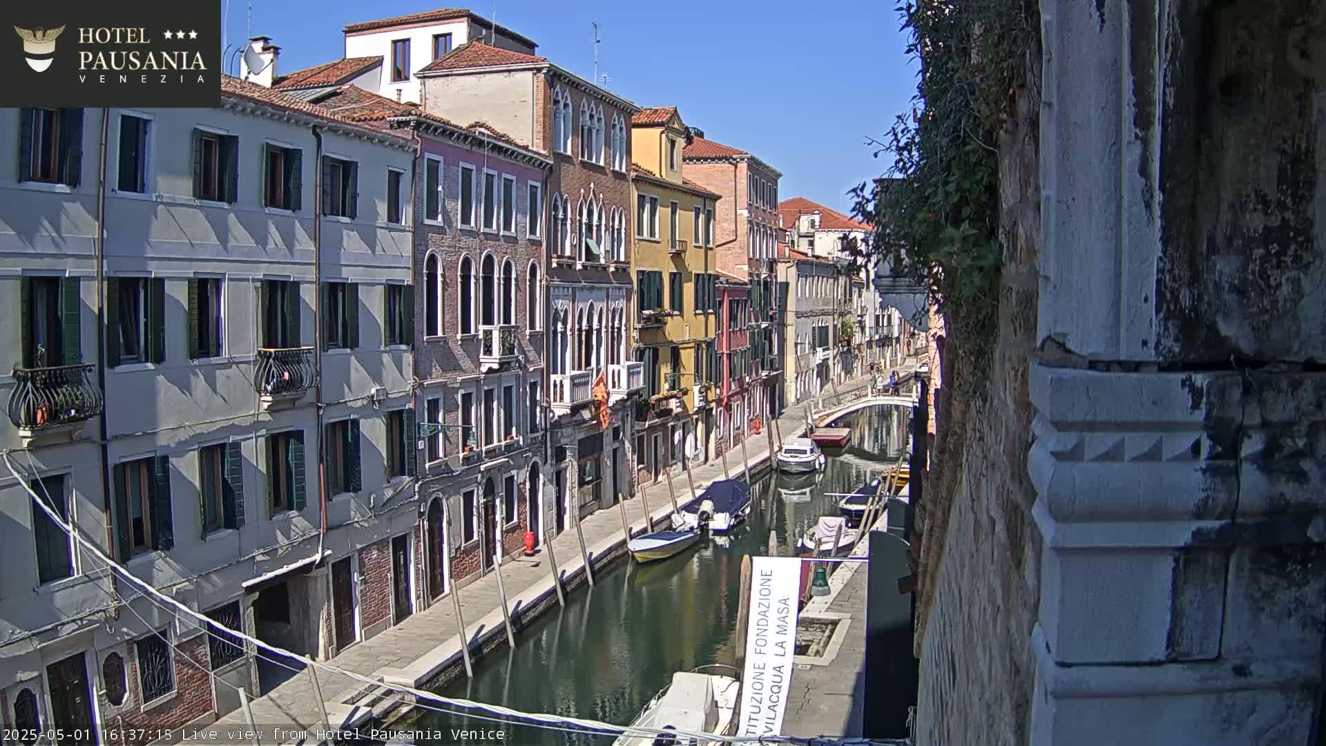 A sunny day in Venice shows a narrow canal lined with colorful buildings, several small boats moored along its banks.