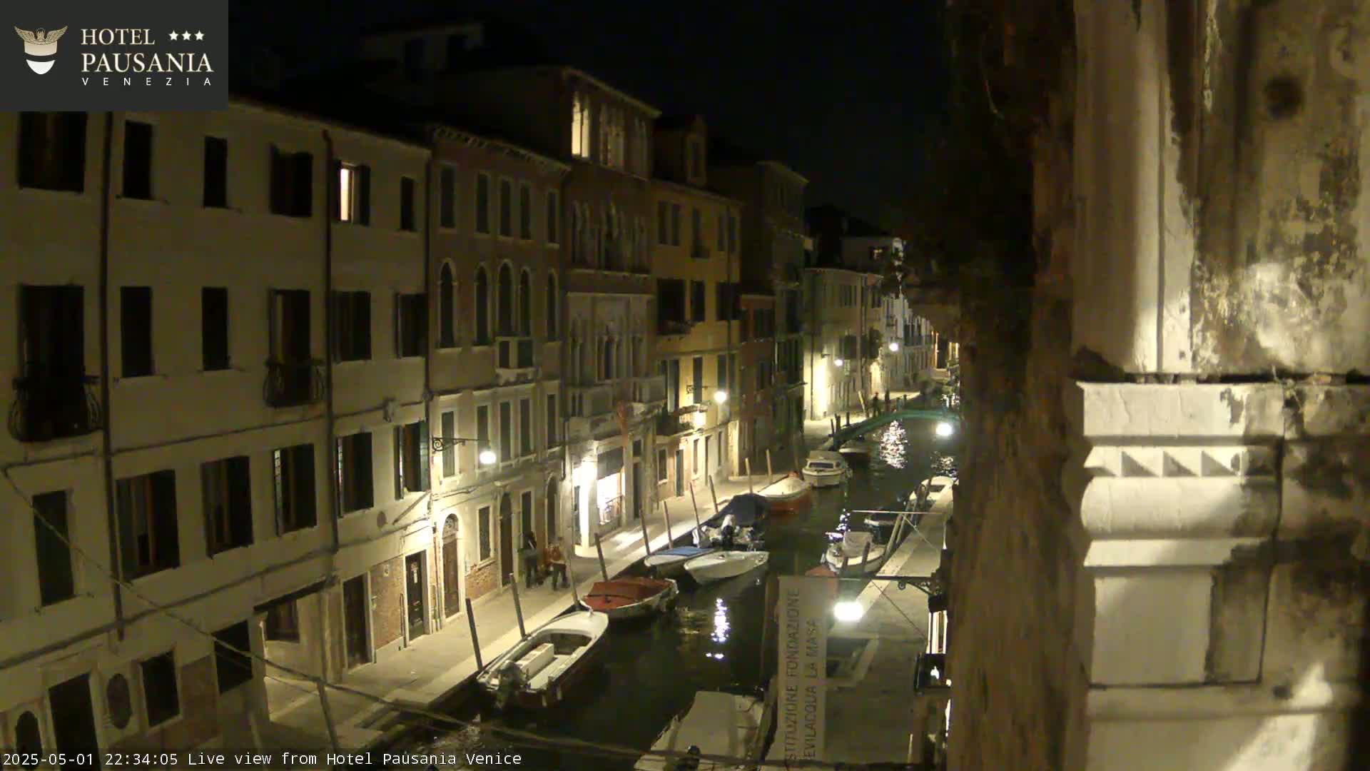 A nighttime view of a narrow canal in Venice, lined with illuminated buildings and several moored boats.