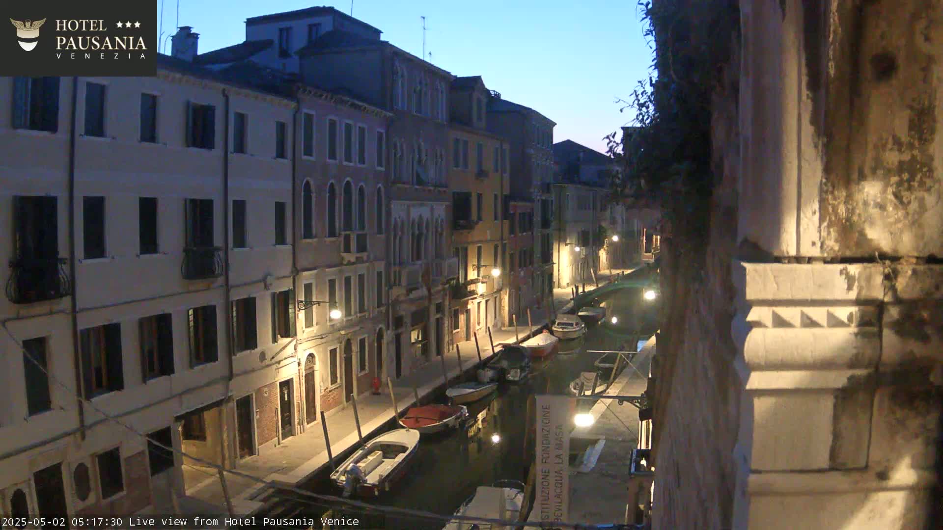Several boats are moored along a narrow canal at twilight, lined with tall, dimly lit buildings.