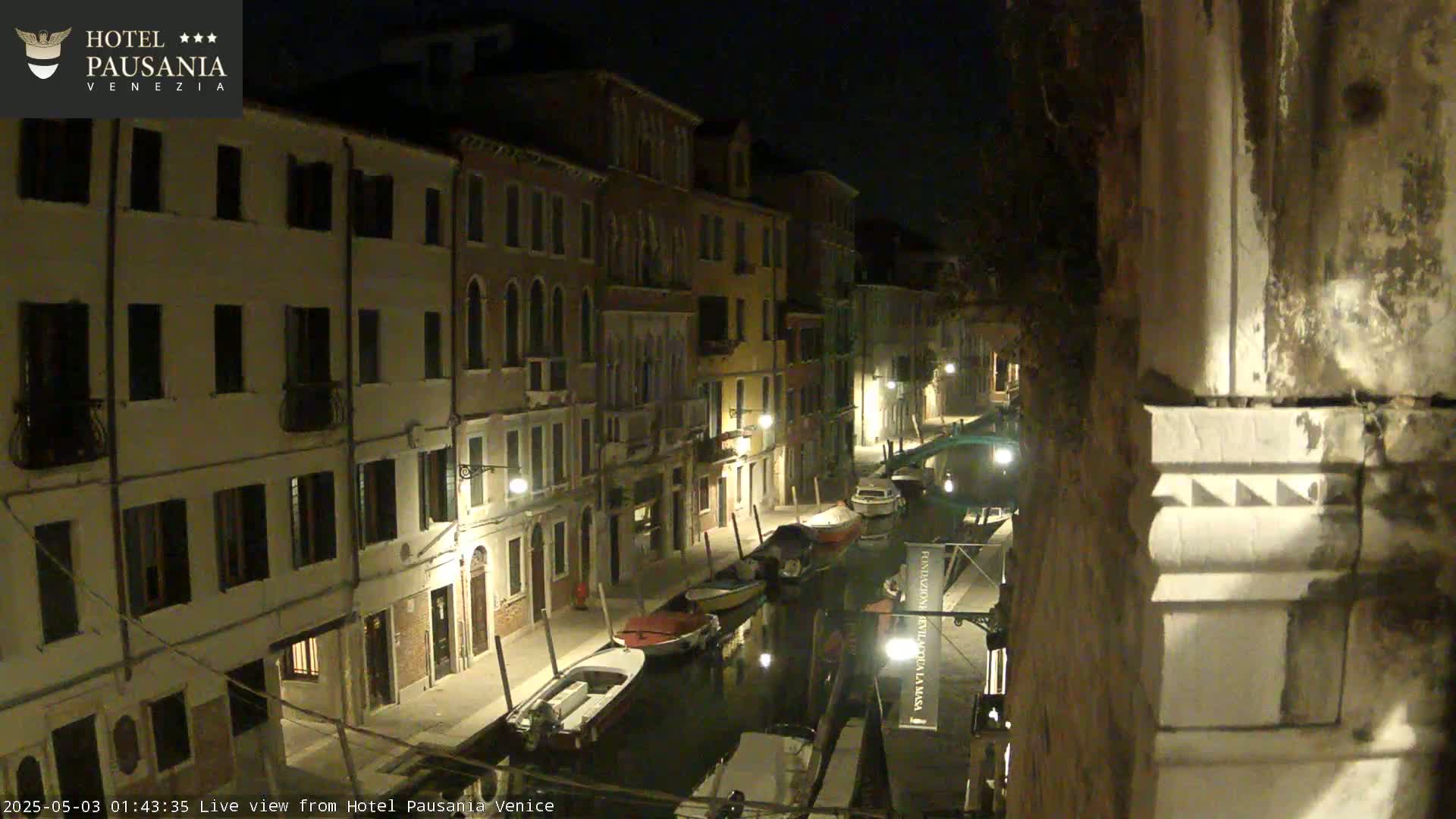 A nighttime view of a narrow canal in Venice, Italy, lined with buildings and several moored boats.
