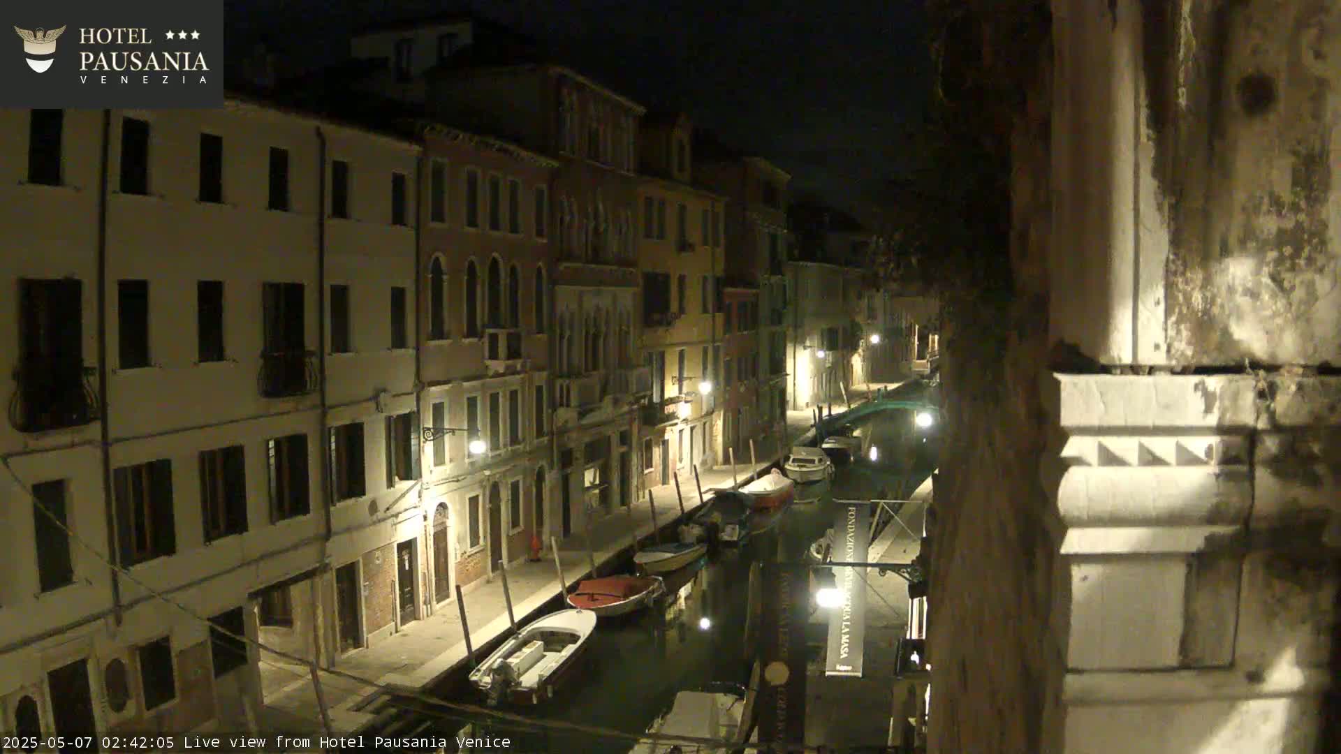 A nighttime view of a canal in Venice, showing several boats moored along the waterway, with illuminated buildings lining the banks.