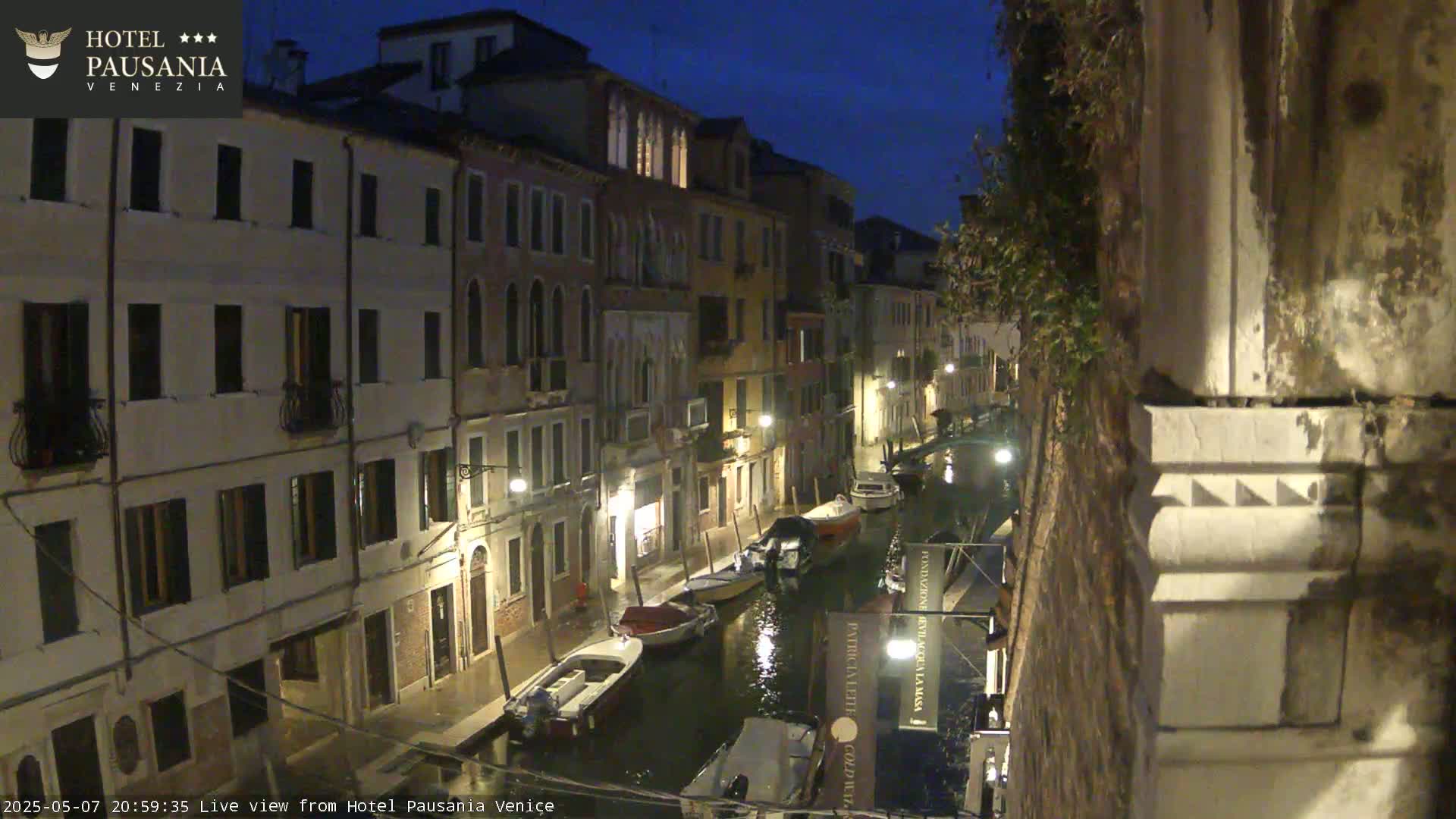 Several boats are moored along a canal at night, flanked by illuminated buildings.