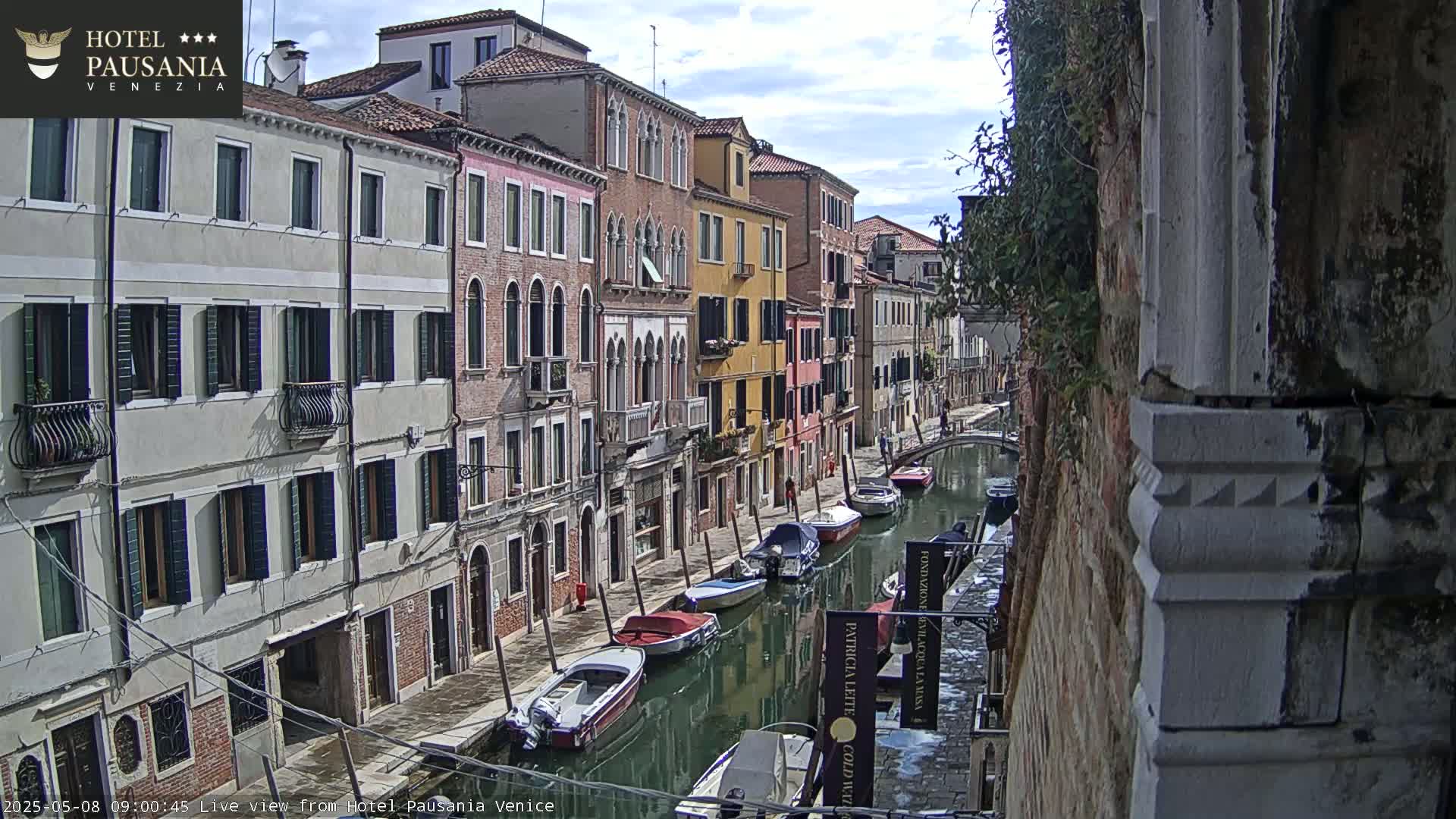A narrow Venetian canal is lined with colorful buildings, several motorboats and small vessels are moored along its sides under a partly cloudy sky.
