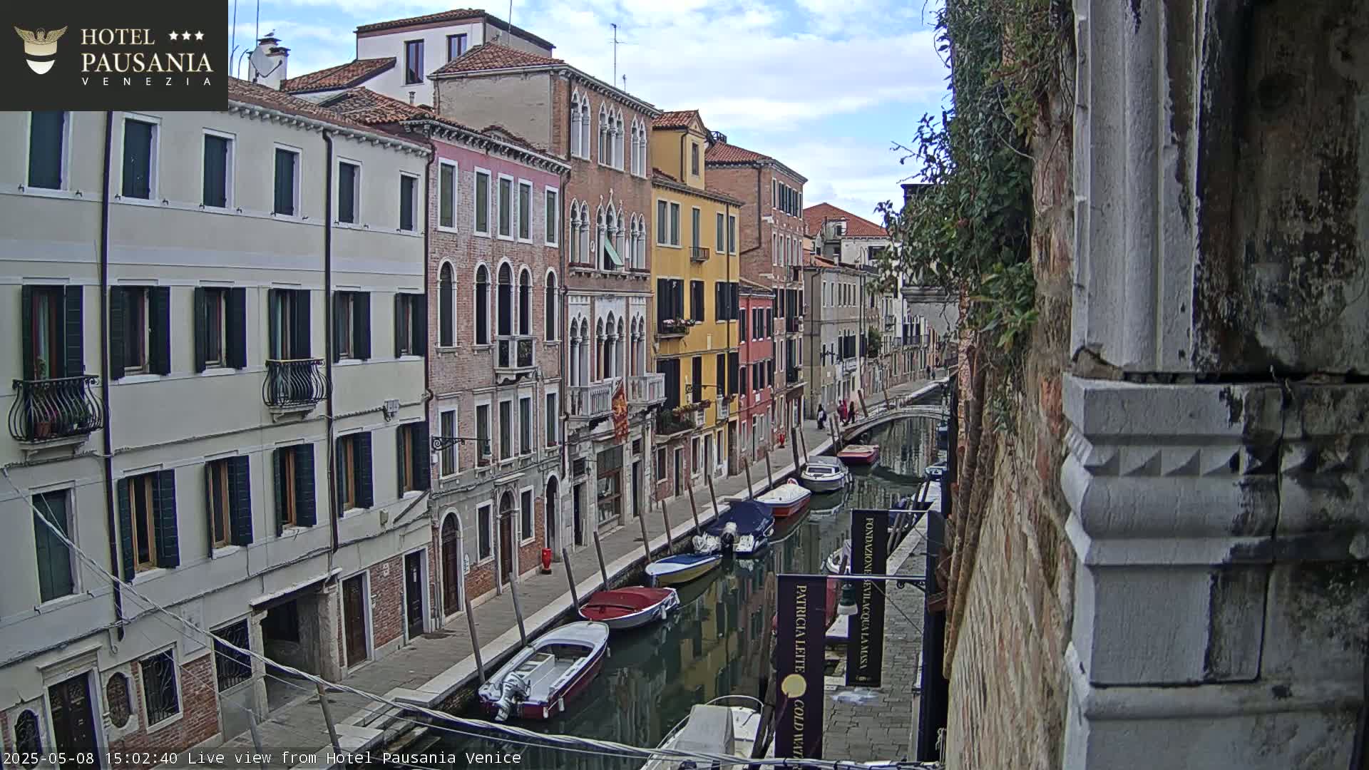 A canal in Venice, under partly cloudy skies, is lined with colorful buildings and several moored boats.