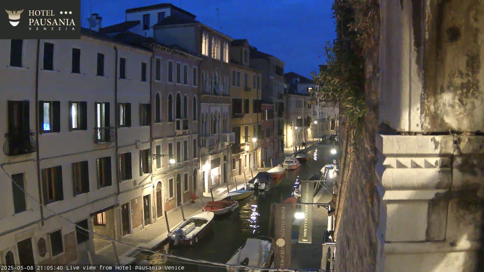 A nighttime view of a narrow canal in Venice, Italy, lined with illuminated buildings, showing several moored boats on the water under a dark blue sky.