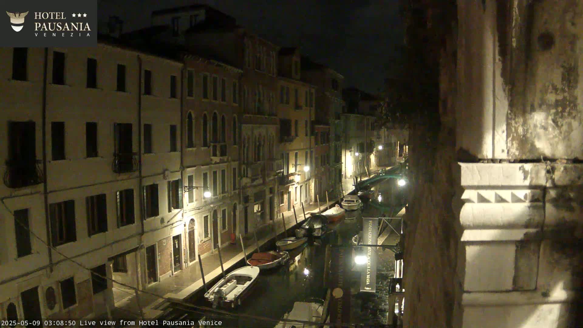 A nighttime view of a narrow canal in Venice, lined with tall buildings, shows several moored boats under dimly lit streets.