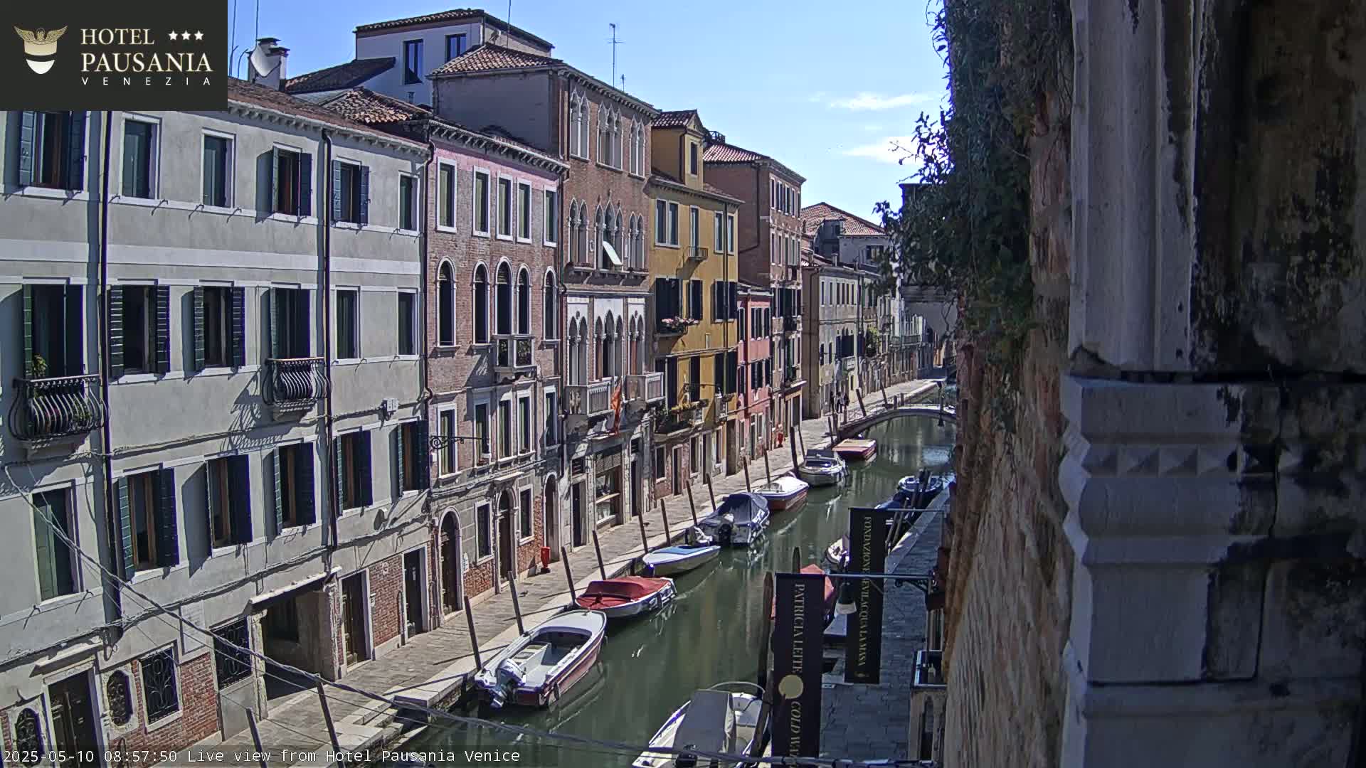 A sunny day in Venice shows a narrow canal lined with colorful buildings and several small motorboats moored along the waterway.