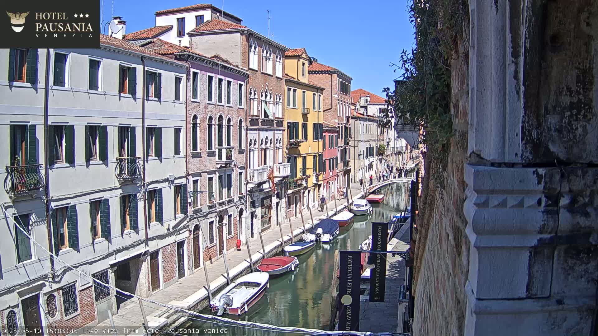 A sunny day in Venice shows a narrow canal lined with colorful buildings and several moored boats.