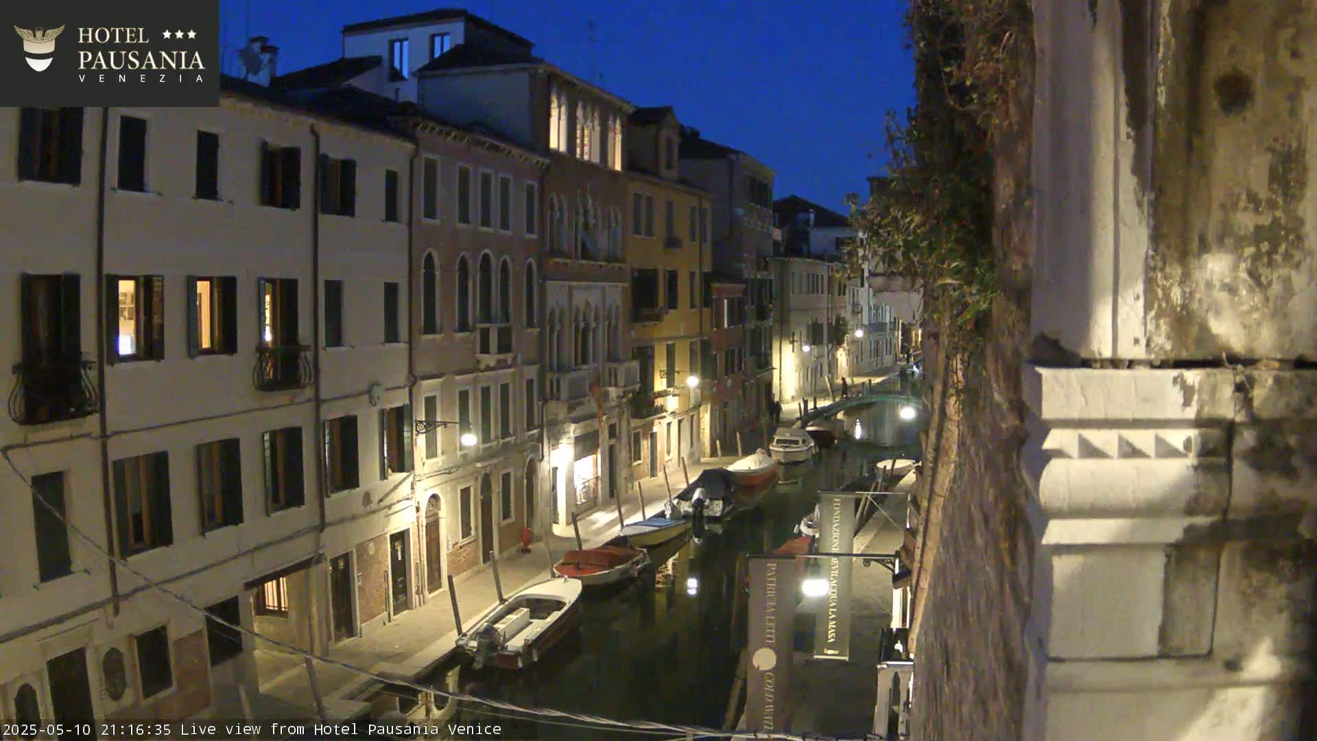 A nighttime view of a narrow canal in Venice, with boats moored along its sides and illuminated buildings lining the waterway.