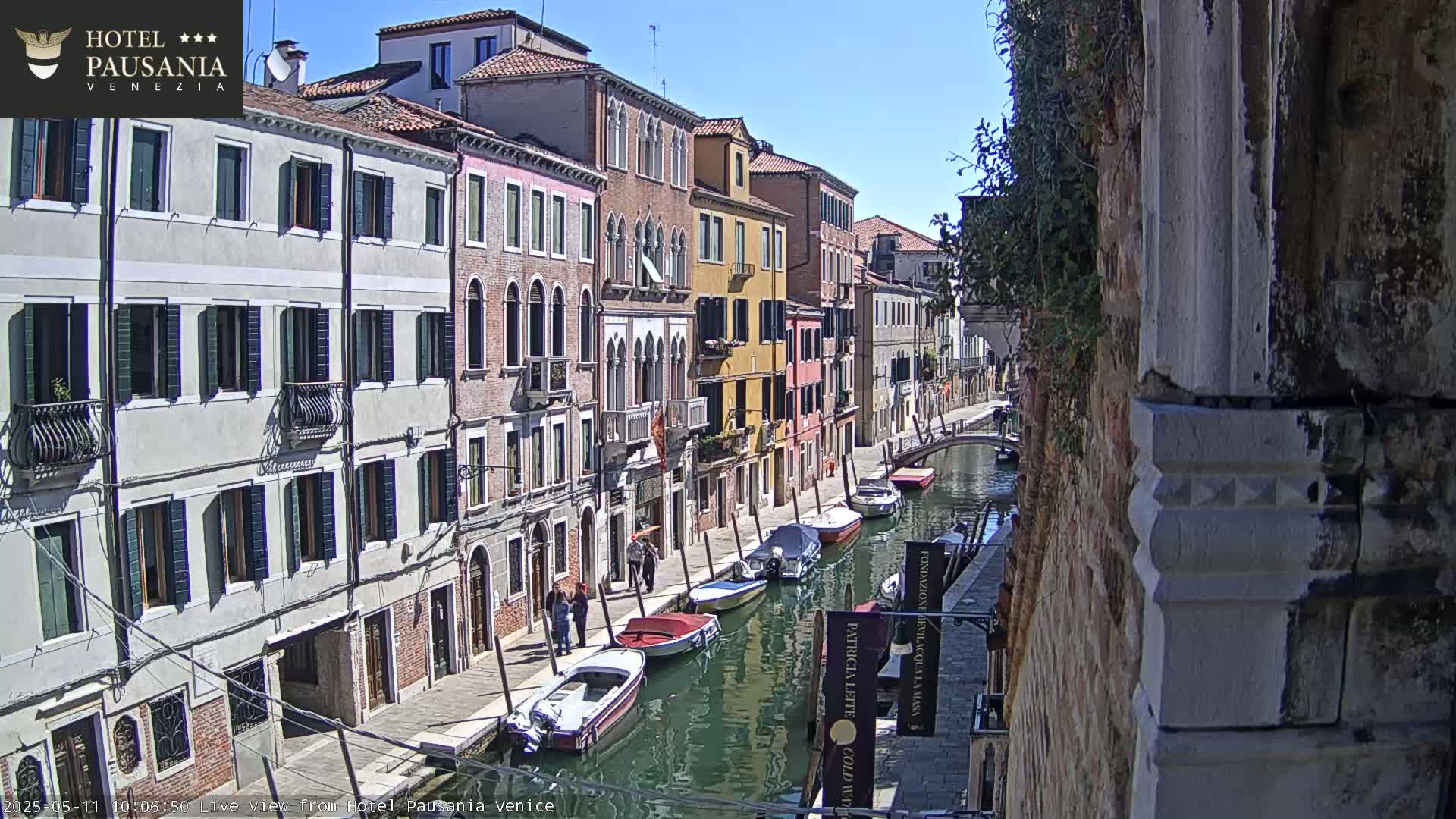 A sunny Venetian canal scene shows several moored boats alongside a narrow waterway bordered by multi-storied buildings.