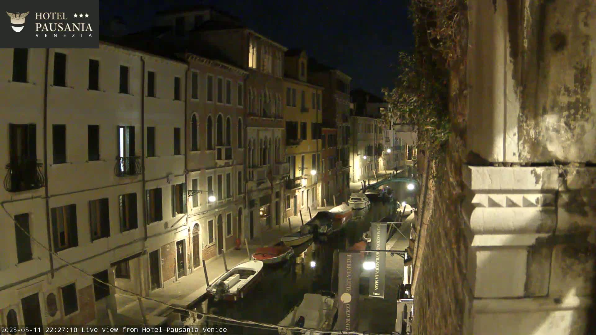 A nighttime view of a narrow canal in Venice, lined with illuminated buildings and several moored boats.