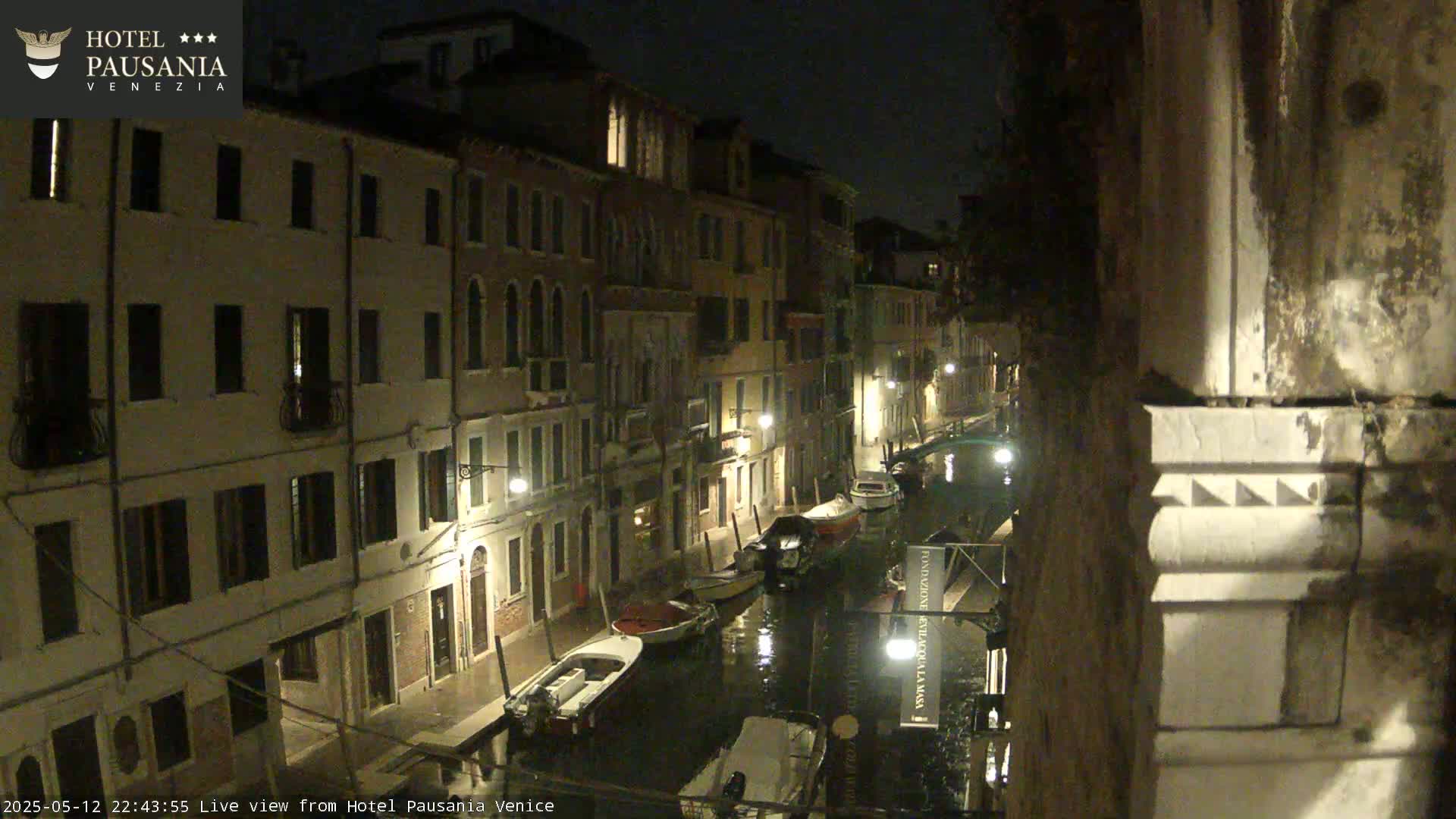 A nighttime view of a narrow canal in Venice, with several boats moored along the sides of buildings illuminated by streetlights.