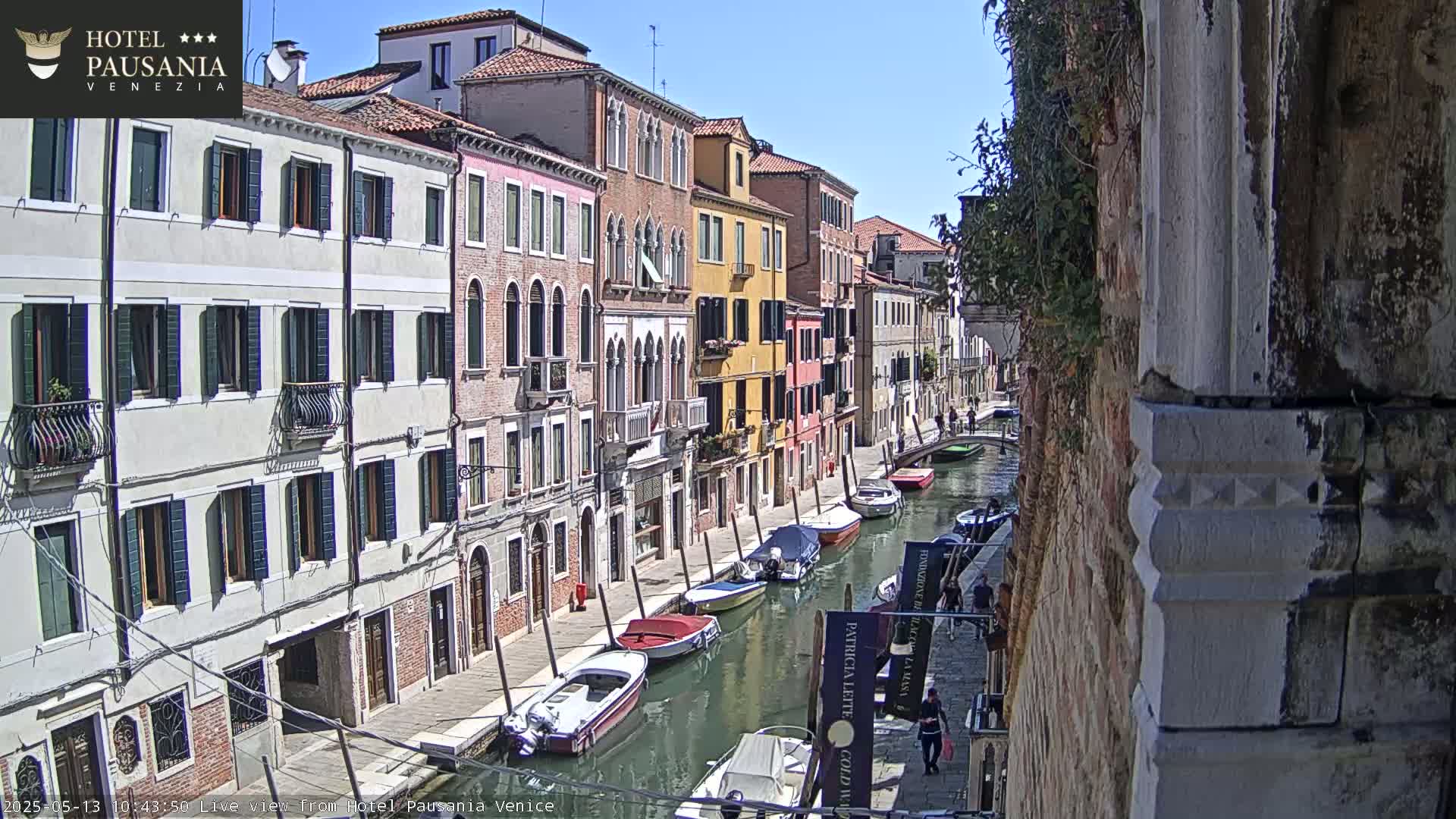 A sunny day in Venice shows a canal lined with colorful buildings and several motorboats moored along the waterway.