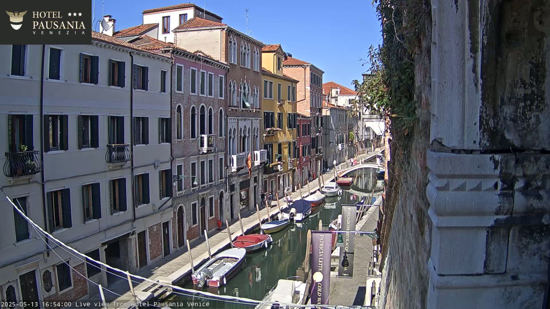 A sunny day in Venice shows a narrow canal lined with colorful buildings and several moored boats.