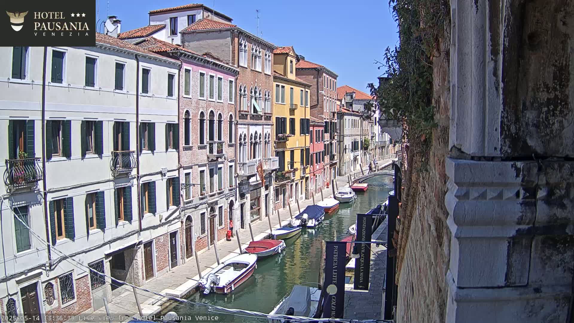 Several boats are moored along a narrow canal in Venice, under a bright sunny sky, beside colorful buildings.