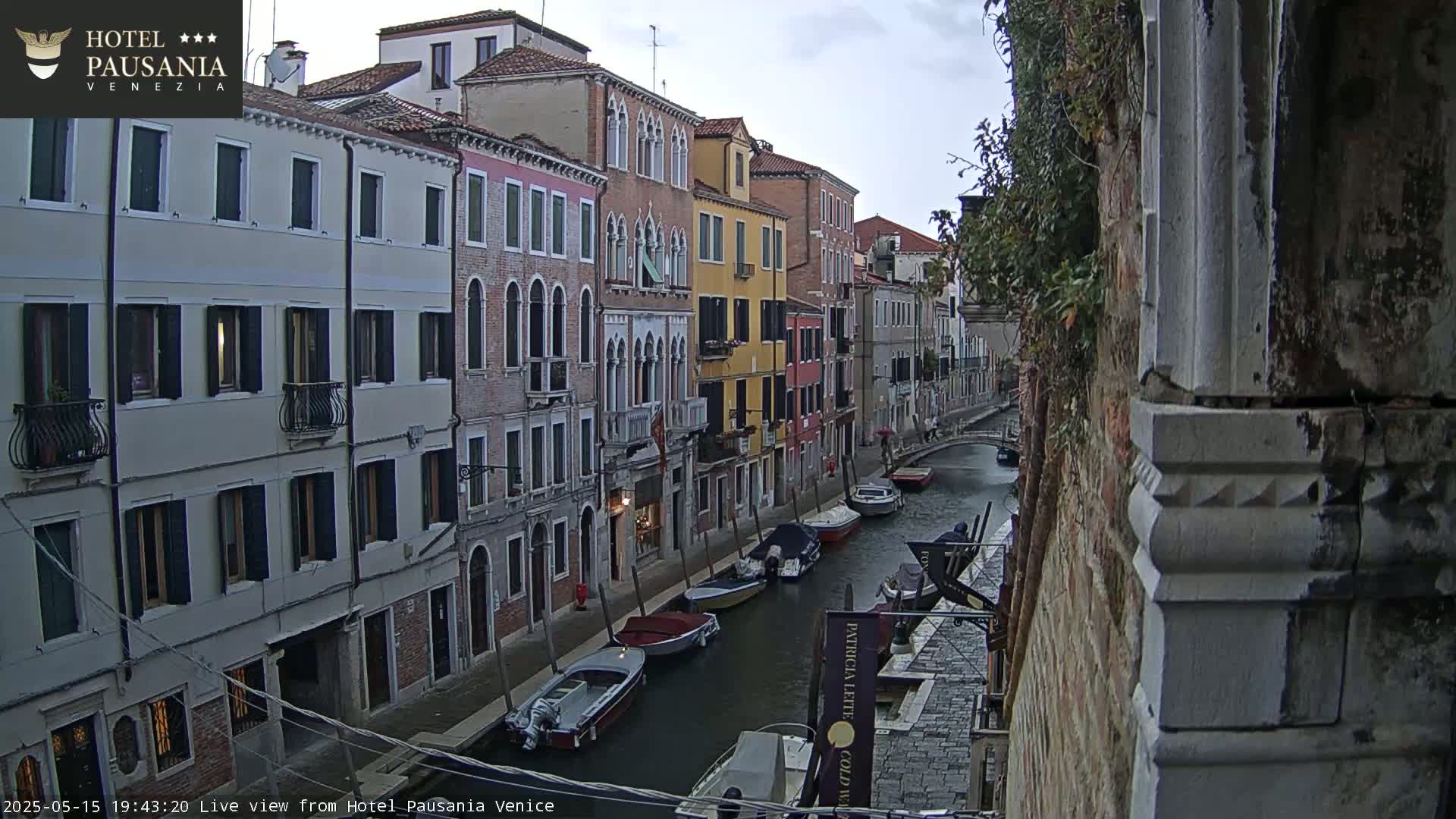 Several boats are moored along a narrow canal in Venice, under an overcast sky, lined with tall, multi-story buildings.
