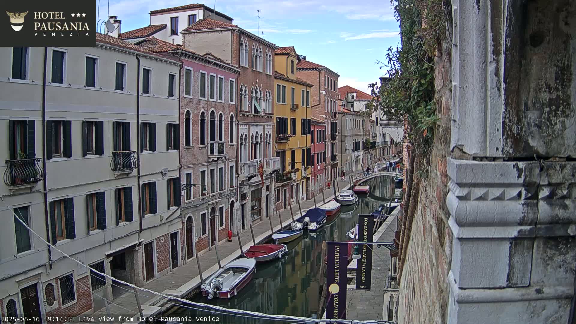 A canal in Venice, lined with colorful buildings, under a partly cloudy sky, contains several moored motorboats and rowboats.