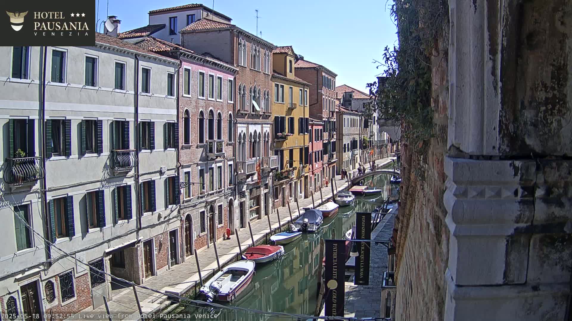 A sunny day in Venice shows a narrow canal lined with colorful buildings and several moored boats.