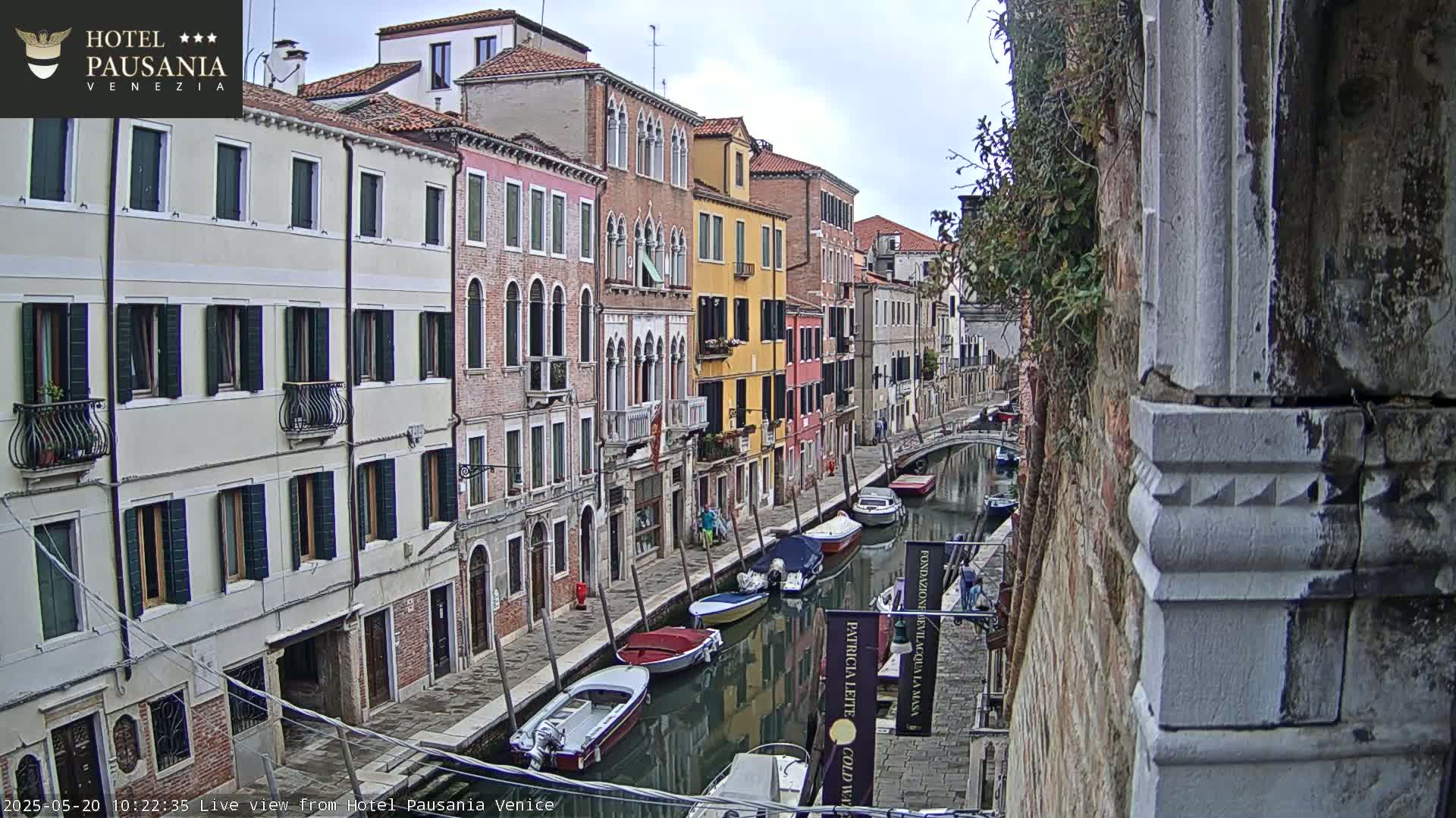 A narrow canal in Venice, under an overcast sky, is lined with colorful buildings and several moored boats.